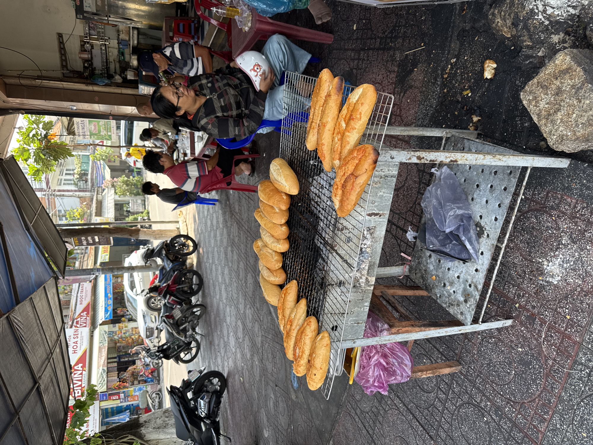 Here is a detailed description of the image:
**General Overview:** The image shows a street scene in what appears to be Bac Nha Trang Ward, Vietnam. The perspective is an elevated, slightly downward angle, capturing a bustling street with vendors, customers, motorcycles, and buildings.
**People:** Several people are visible, mostly seated and interacting with a roadside food stall. One person is wearing a plaid shirt and glasses, seemingly engaged in conversation. Another person is wearing a blue shirt with a helmet on the chair.
**Subjects:** The main subjects are the food stall and the long loafs of bread being grilled. The bread looks toasted.
**Objects:** The image includes motorcycles parked along the sidewalk, food stalls with metal grilling racks, plastic chairs, rocks, and the bags.
**Actions/Interactions:** People are seated, presumably eating or conversing. The vendor appears to be preparing or serving food. Motorcyclists are moving along the street.
**Environment:** The location appears to be a busy street in Bac Nha Trang Ward. Buildings line the street, with signs in Vietnamese visible. The sidewalk has a patterned design.
**Essential Details:**
* **Location:** Bac Nha Trang Ward, Vietnam.
* **Activity:** A roadside food stall is in operation.
* **Focus:** Toasted bread (banh mi).
* **Environment:** Outdoor, urban street scene.
* **Transportation:** Motorcycles parked along the street.