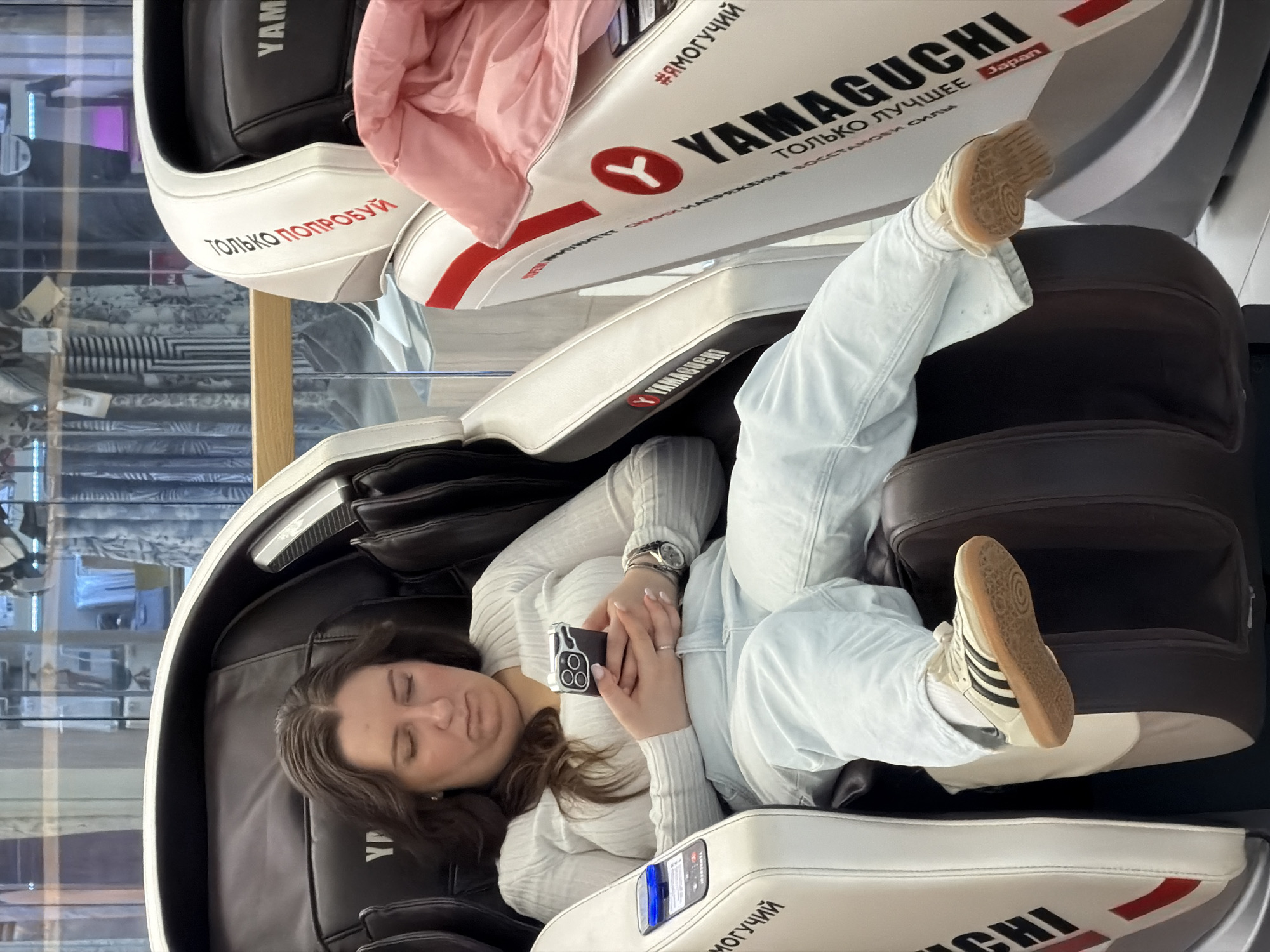 The scene depicts a woman relaxing in a massage chair in a store, with the brand name "Yamaguchi" prominently displayed. The chair is white with black and red accents, featuring text in Russian, including phrases like "#ЯМОГУЧИЙ" (which translates to "#IAMPOWERFUL"), "ТОЛЬКО ЛУЧШЕЕ" ("ONLY THE BEST"), and "ТОЛЬКО ПОПРОБУЙ" ("JUST TRY"). The tag "Japan" is also visible on the chair.
The woman, dressed in a light-colored sweater and jeans, is lying back in the chair with her eyes closed, holding her phone. She is wearing a watch on her left wrist and a ring on her left ring finger. Her legs are extended, and her feet are wearing white sneakers with black stripes.
The background appears to show a shop window with shelves displaying various items, possibly clothing. Given the Russian text and the store setting, it is plausible this scene is taking place in Moscow, Russia. The environment suggests a consumer-oriented space where people can test or purchase massage chairs.