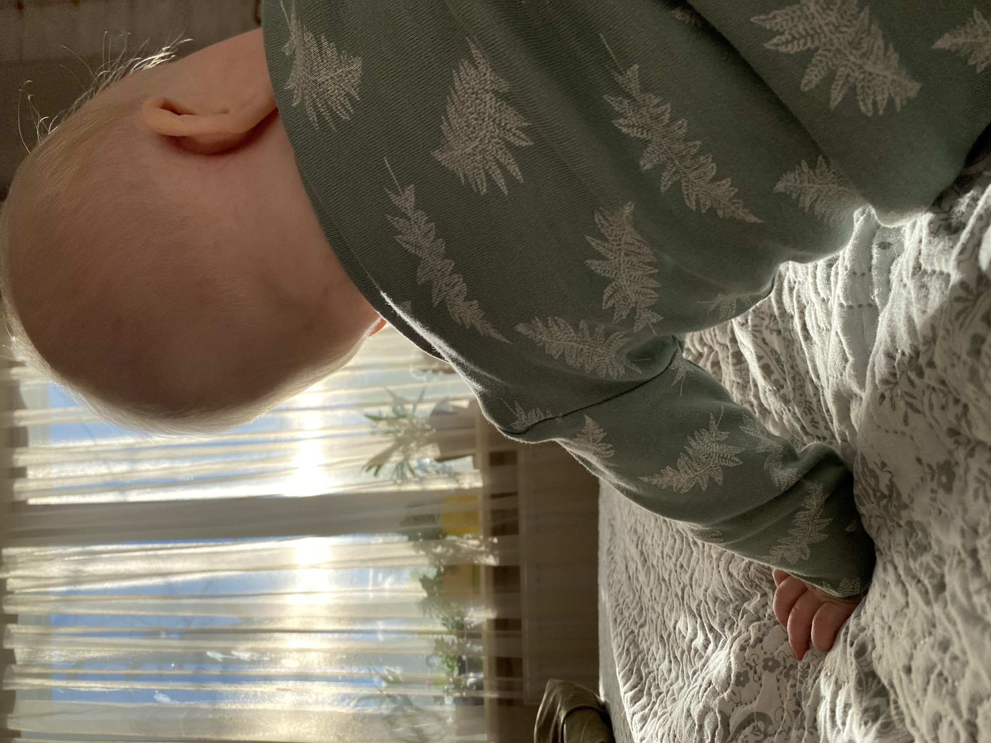 A close-up shot depicts a fair-skinned baby, primarily showing the back of its head and ear, positioned near a window. The baby is wearing a long-sleeved garment of light green color with a white fern-like design. The baby's hand is visible, slightly buried within a textured light gray bedspread. Sunlight streams through a window covered with light, translucent curtains. The location in the city of Фоки, Russia is indeterminate from the visual information.
