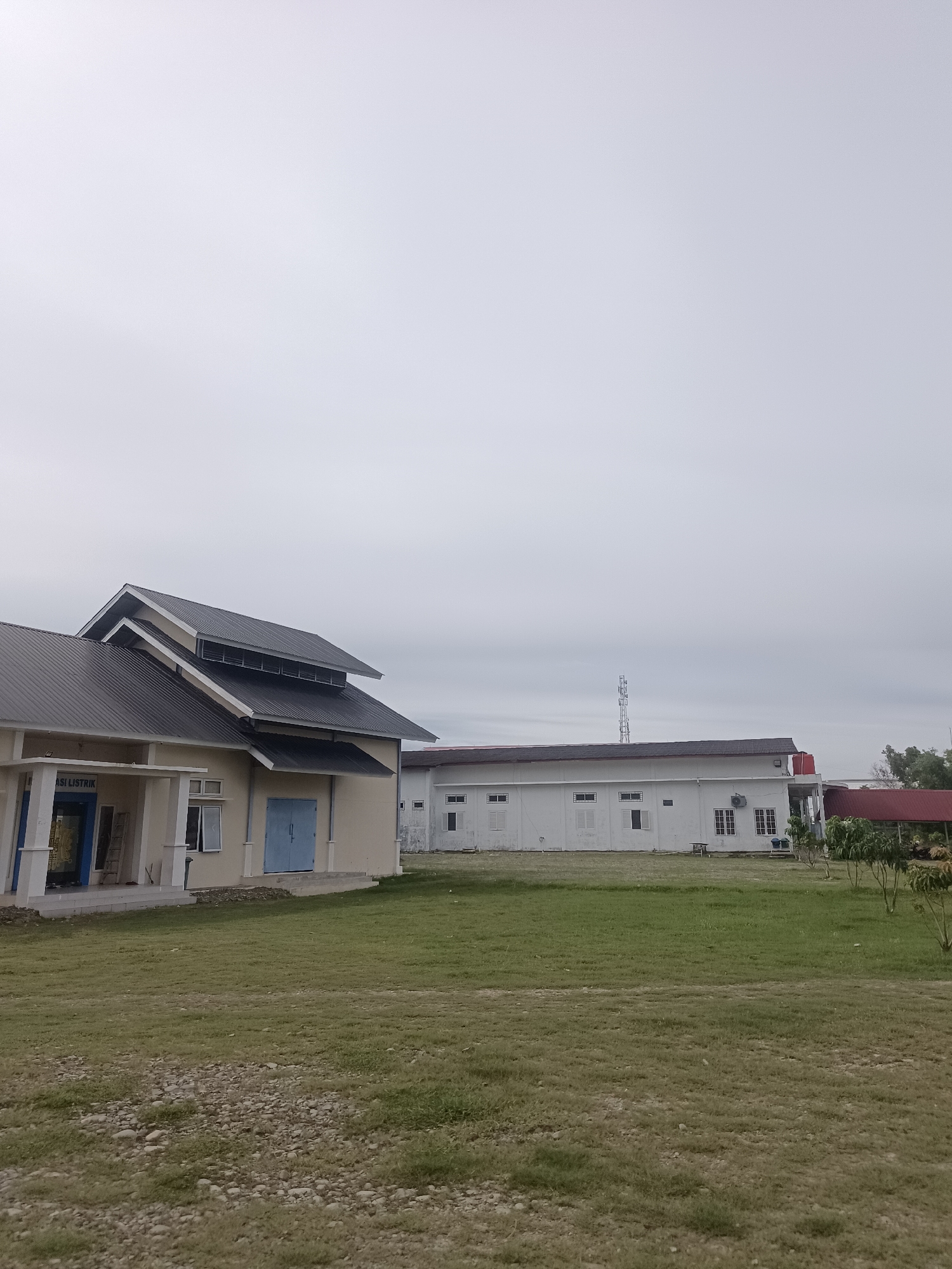 This is a vertical, medium-shot photograph taken from a low angle, looking up at a cluster of buildings under an overcast sky. The scene appears to be in Mesjid Reuntoh, Indonesia.
In the foreground, a patch of dry, patchy grass with scattered small stones covers the ground.
Dominating the left side of the frame is a light-colored building with a dark, sloped metal roof. The building has a wide overhang supported by columns. Visible text above a doorway on this building reads "ASI LISTRIK." A light blue double door is present on the side of this building. Two windows are visible, one open. A ladder leans against the building near the entrance.
To the right of the first building is a long, white, rectangular building with a dark roof. This building features numerous windows, some of which are open. An air conditioning unit is mounted on the exterior wall, and a water tank with a red cap is visible near the right end. A cell tower is visible in the distance, behind this building.
In the far right of the frame, a smaller building with a red roof is partially visible, partially obscured by green foliage and the branches of trees.
The sky is uniformly gray and cloudy, providing diffused, flat lighting across the entire scene. No people are visible in the photograph.