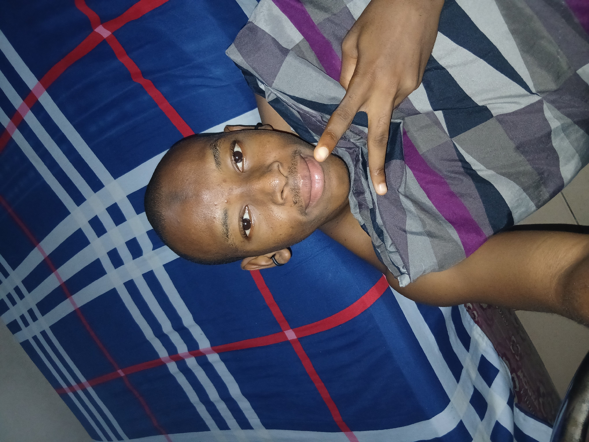 A young Black male is lying on a bed with a blue and white checkered blanket, with a red stripe running diagonally across it. He is looking at the camera and holding up two fingers in a peace sign. He is wearing a black earring in his left ear and has a mustache. He is partially covered by a gray, purple, and black checkered blanket. The scene appears to be indoors, likely in a bedroom, in Lagos, Nigeria.