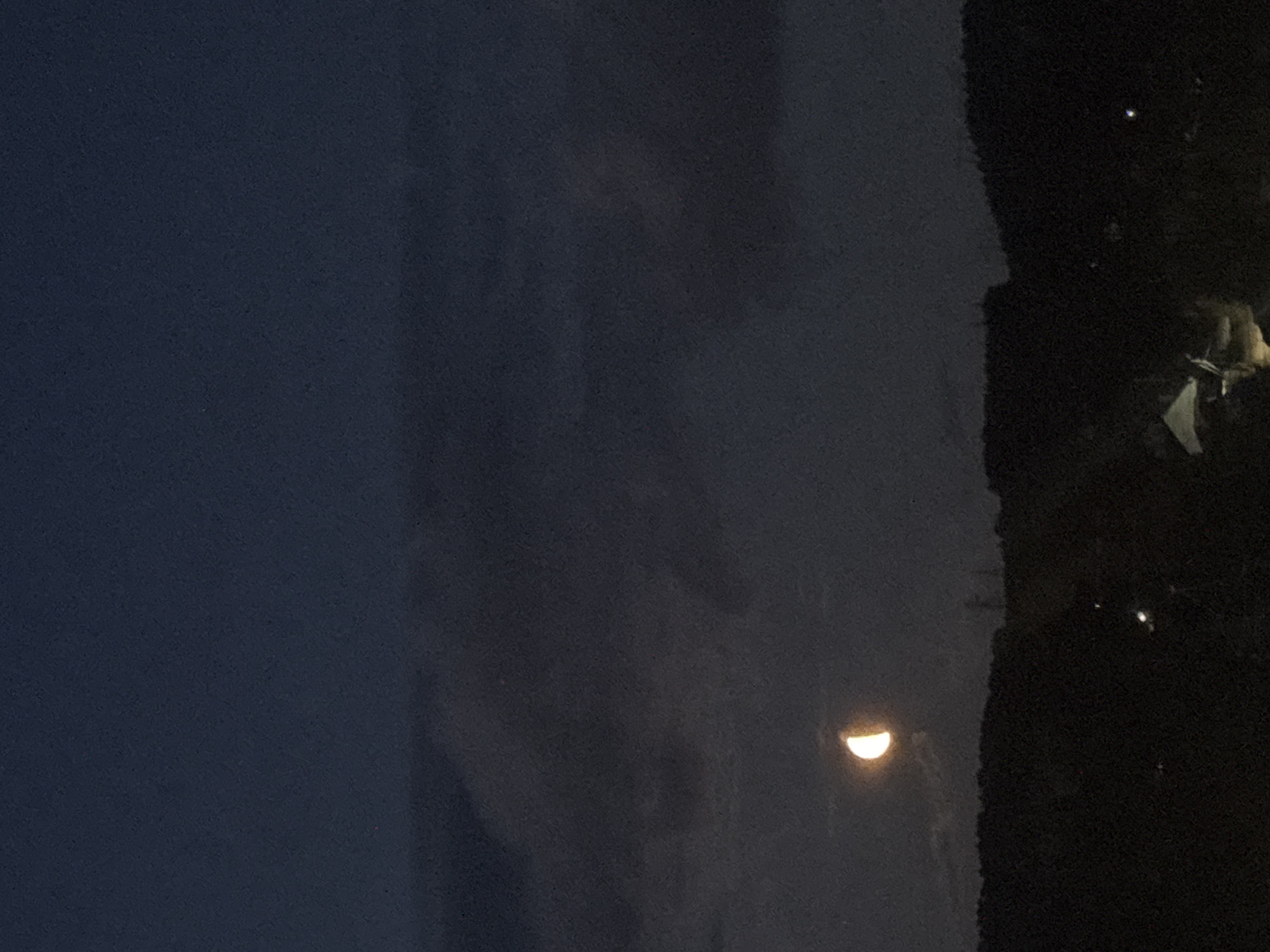 A nighttime scene with a half-moon visible in the sky. To the right, a dark area suggests the presence of trees. In the upper-right corner, there are a couple of bright lights, possibly from streetlamps or distant buildings. The scene is likely in an outdoor setting, given the presence of the moon and trees. There's no information in the image to confirm the location as Chaikovsky, Russia.