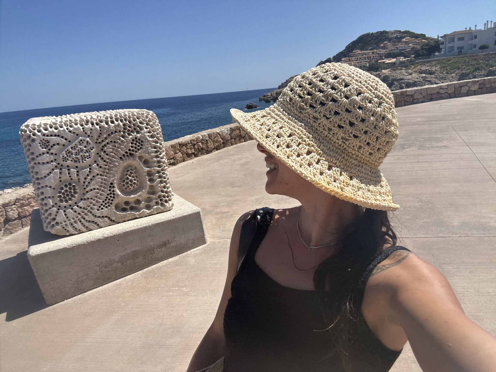 In Cala Lliteres, Spain, on a bright, sunny day, a woman is captured in a selfie, posing beside a stone sculpture. The sculpture, a textured white rectangular block with intricate, lace-like perforations and circular patterns, sits on a beige concrete pedestal. The woman, wearing a wide-brimmed woven straw hat and a black tank top, turns her head to the left, her profile visible as she smiles. A delicate silver necklace adorns her neck. Her left arm, partially visible, displays a tattoo on her shoulder. Behind her, the deep blue Mediterranean Sea stretches out, meeting a clear, light blue sky. A stone wall, constructed from rough, irregular blocks, runs along the coastline. In the distance, a hillside is dotted with white buildings, a cluster of houses nestled amongst the terrain. The ground in the foreground is a light beige paved surface, with distinct lines marking sections of the pavement. The overall scene depicts a woman enjoying a scenic coastal view.