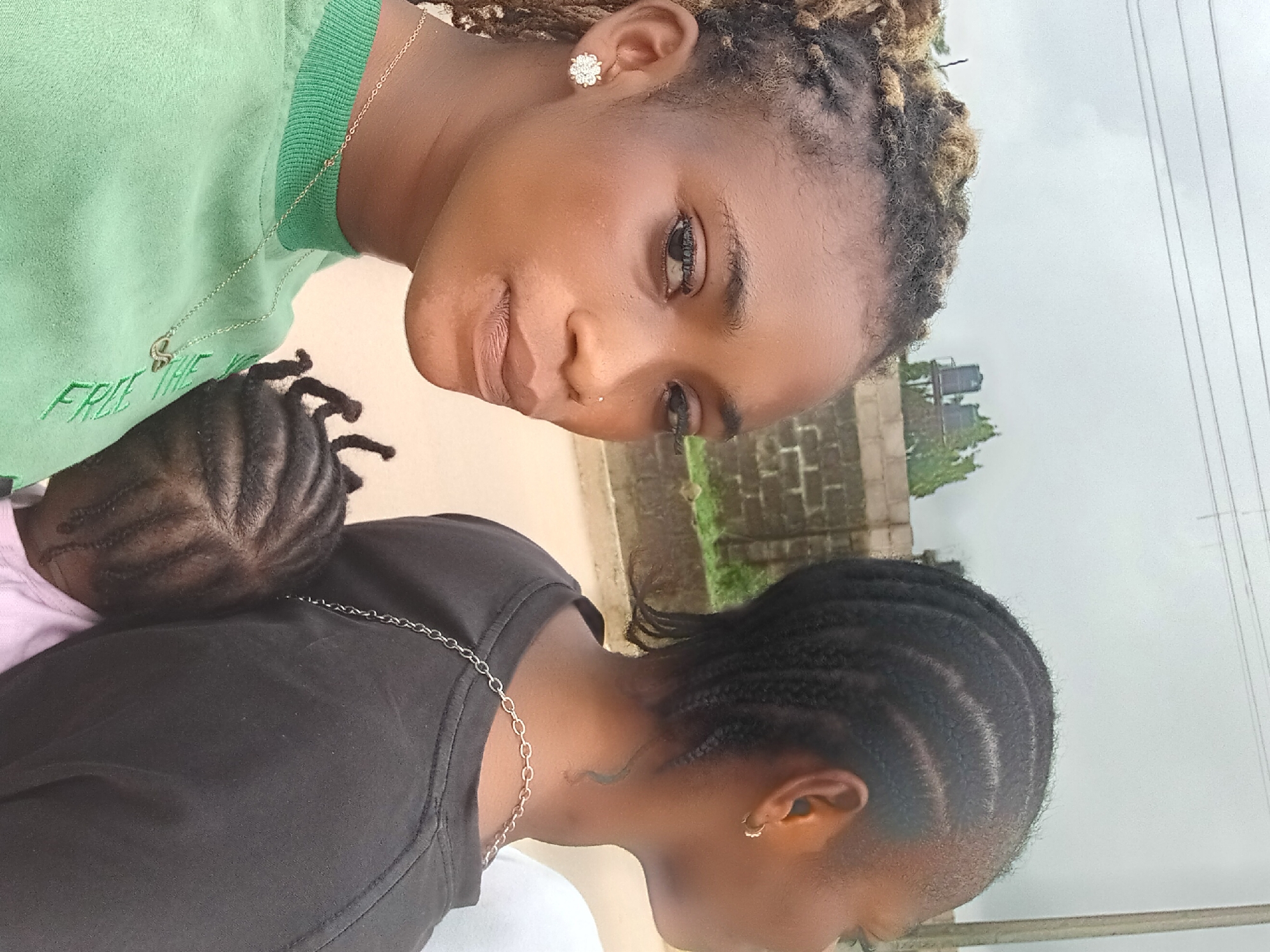 A medium shot of two Black women, one in a green shirt and the other in a black shirt, with a child who is resting on the shoulder of the woman wearing the black shirt. The woman in the green shirt faces the camera and has dark eyes, and wears an earring shaped like a flower. The text "FREE THE" is embroidered in green on the green shirt. A gold chain hangs around her neck. Her hair appears to be in locs. The other woman's face is not visible, but it appears that her hair is braided, and she is also wearing a gold chain. The child's hair is braided. The background shows a concrete building structure and what appears to be utility poles. Based on the appearance of the people and the vegetation, this image might have been taken in the city of Shagamu, Nigeria.