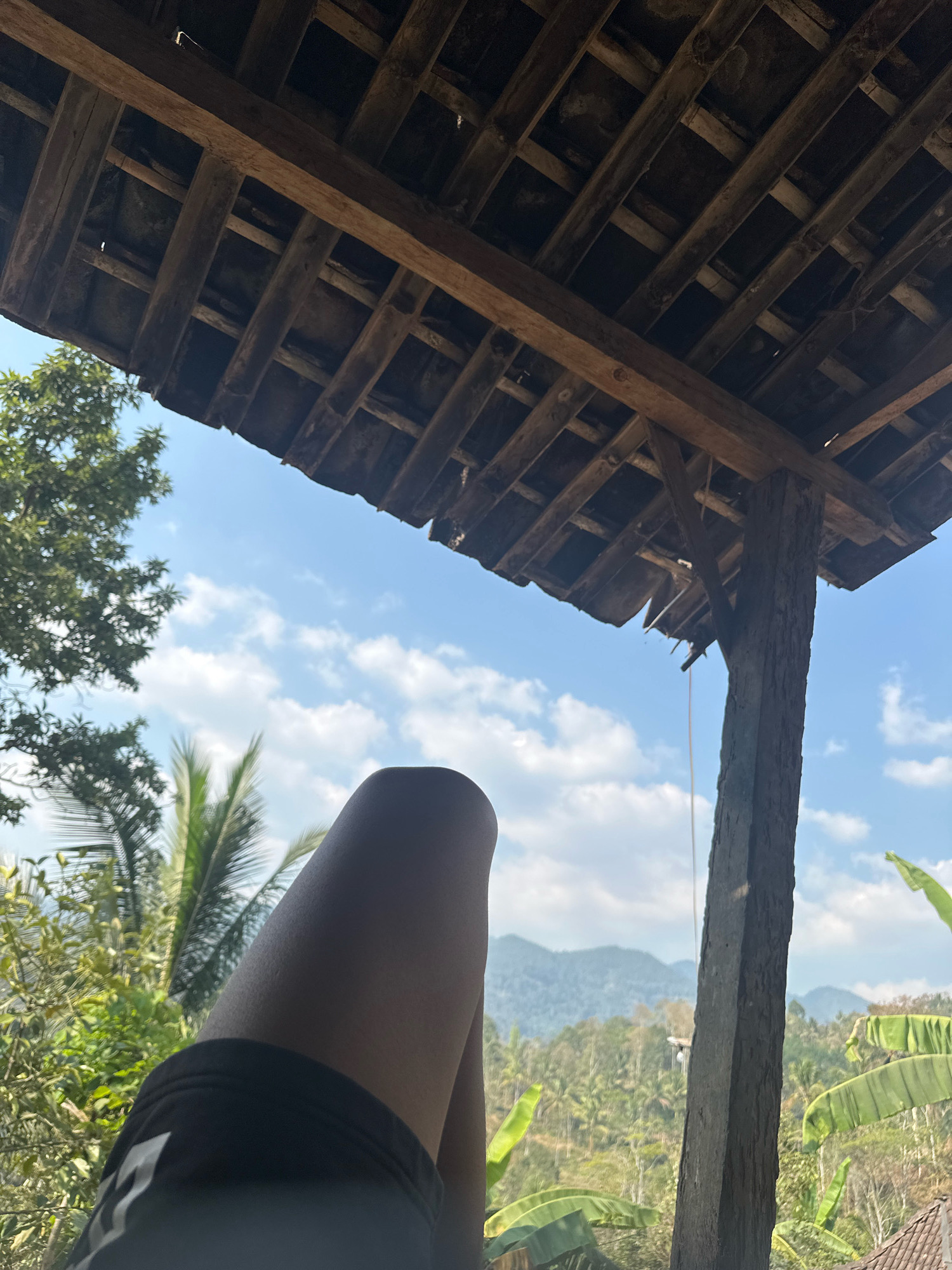 The image shows a close-up of a person's legs and shorts, with their upper body cropped out of the frame. The legs are bent at the knee, and the person is wearing black shorts with white accents. The background is a lush green landscape with palm trees and mountains, under a bright blue sky with scattered clouds. This scene is captured from under a rustic wooden structure, possibly a veranda or open-air pavilion, with exposed wooden beams and rafters visible. The overall impression is one of relaxation and enjoying a scenic view, likely in a tropical or subtropical environment. The location is identified as Talun, Indonesia.