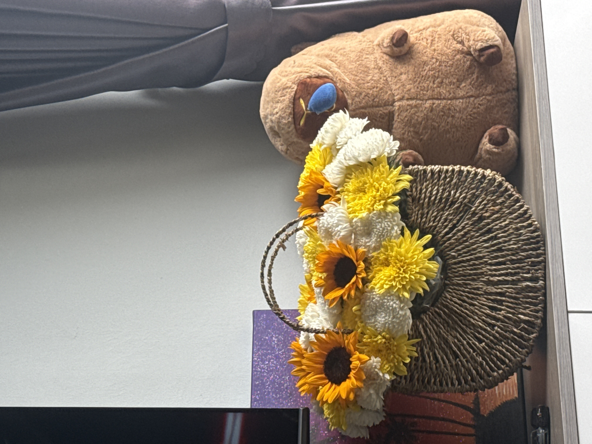 A still life arrangement is presented on a light-colored shelf in front of a neutral-toned wall and dark grey curtains. Dominating the left side of the arrangement is a woven basket filled with a vibrant bouquet of yellow sunflowers and white and yellow chrysanthemums. To the right of the basket, a large, plush, tan-colored stuffed toy with a blue tongue and brown feet sits. Behind the flowers, a textured purple rectangular object is visible, and to its left, a dark rectangular object, possibly a television screen, is partially seen. The overall impression is a decorative display within an indoor setting. The image does not provide any specific geographical information to determine its location within Nha Trang, Vietnam.