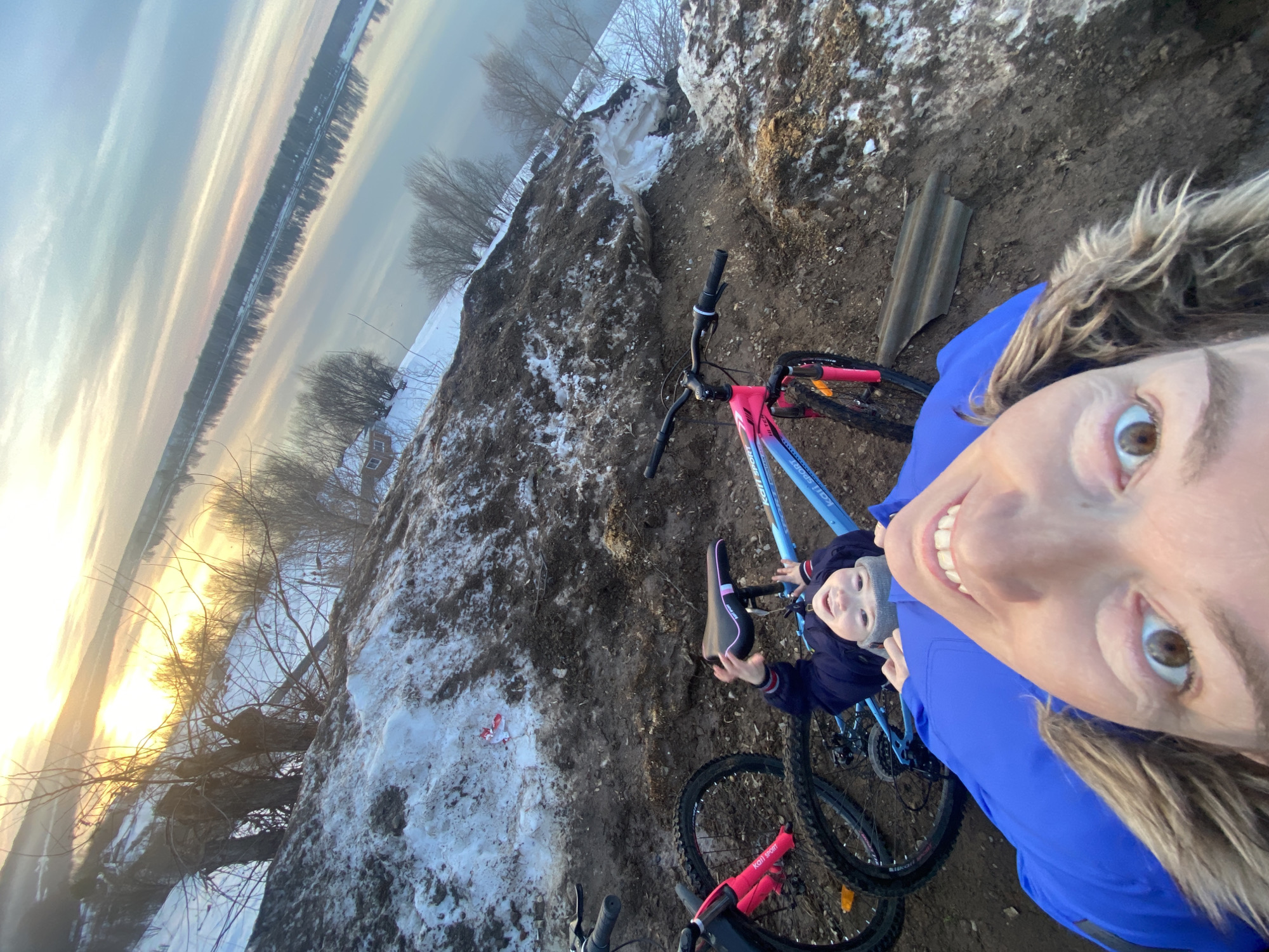 A high-angle selfie photograph captures an adult and a child outdoors on a muddy, snow-covered embankment near a wide body of water in Ольховка, Russia. In the immediate foreground, an adult, likely female, with blonde-brown hair and wearing a bright blue jacket with a fur-trimmed hood, smiles broadly at the camera. Her right eye is wide open, revealing a brown iris. Directly behind the adult, a young child, approximately 3-7 years old, is lying on their back on the dark brown muddy ground, looking up at the camera with a smile. The child wears a dark blue jacket and a grey knitted beanie, with one arm raised.
Two bicycles are visible on the ground. To the left of the child, a light blue mountain bicycle with pink accents on the seat, forks, and handlebars, labeled "Kari Sport," rests on its side. A second, partially visible bicycle with a dark frame and pink front fork is situated to the far left. The ground consists of uneven dark brown mud, interspersed with patches of melting white snow. A small piece of red and white debris is visible within a snow patch near the light blue bicycle. Further to the right, a weathered piece of corrugated roofing material lies on the mud.
The background features a wide, calm body of water, likely a river, reflecting the horizon. The sky exhibits warm orange and yellow hues near the horizon, indicative of either sunrise or sunset, transitioning to lighter blue and grey clouds overhead. Bare deciduous trees and shrubs populate the muddy slope and line the distant bank of the water. A small, light-colored building with a dark roof is faintly discernible on the far bank, partially obscured by trees.