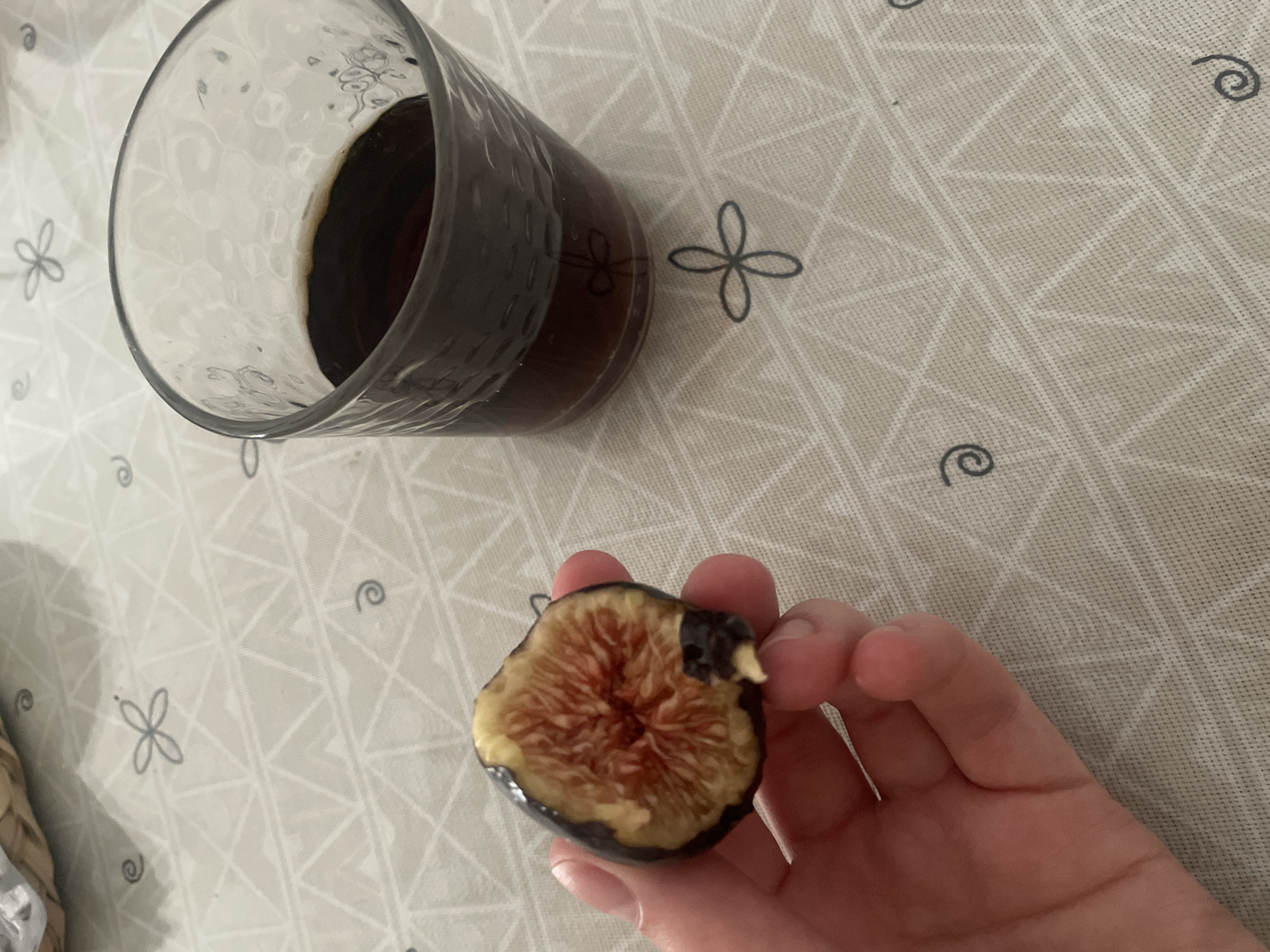 A hand is holding a halved fig, revealing its reddish-pink, seedy interior. To the right of the fig is a clear glass filled with a dark liquid, possibly coffee or a dark beverage. The glass has a textured surface. Both the fig and the glass are resting on a light-colored tablecloth with a pattern of geometric shapes and small, stylized floral or swirled designs. The overall scene is casual and appears to be indoors. The location in the city of Atarfe, Spain, is not determinable from the image.