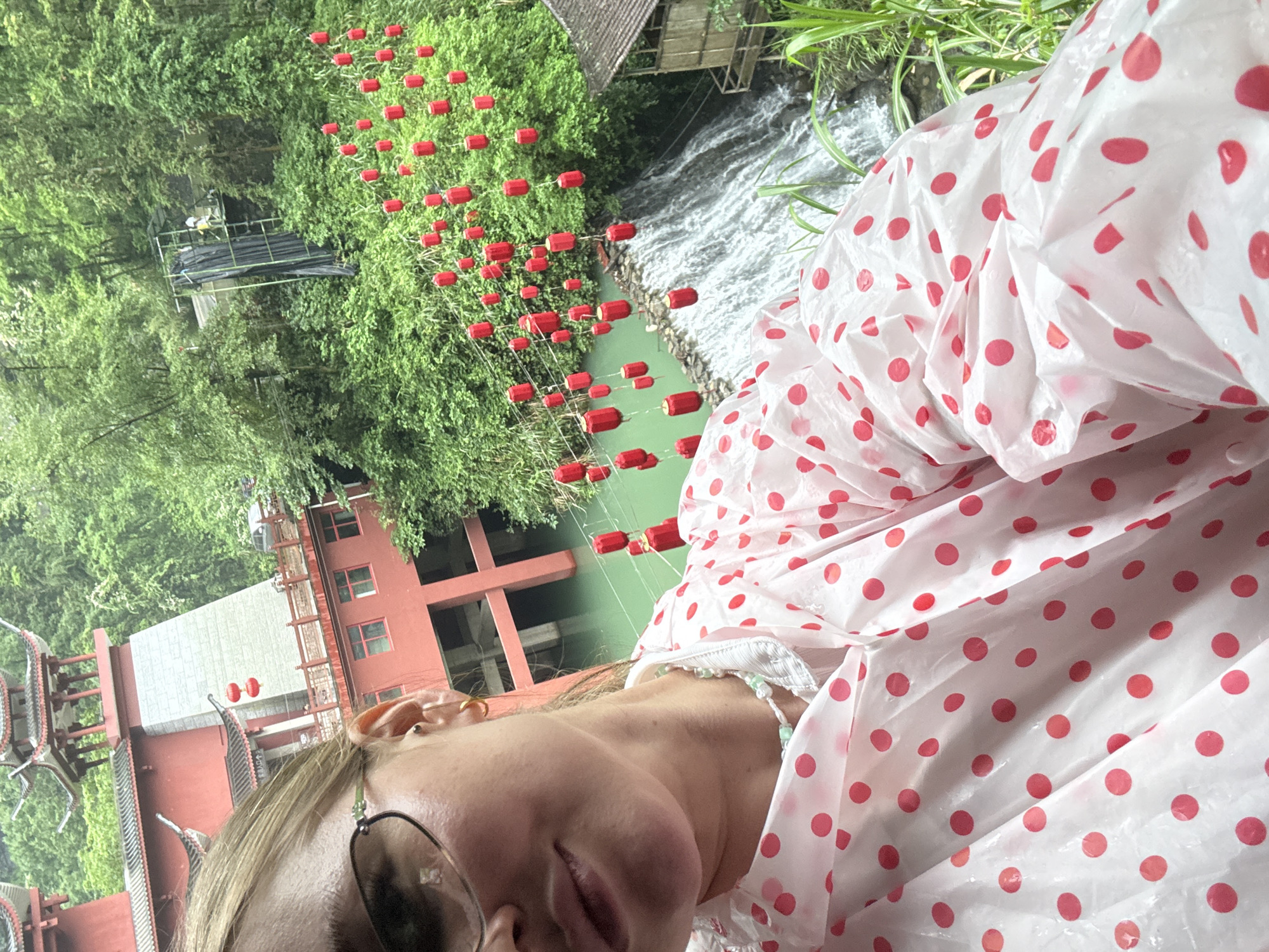 A slightly blurry, canted, close-up, outdoor shot shows a person partially visible in the lower portion of the frame, wearing a white poncho with red polka dots.
In the background, there is a river spanned by numerous red lanterns strung across. Beyond the river, there is a forest. To the left is a traditional Chinese building with a red exterior.
This scene is most likely in Yongding District, Zhangjiajie, Hunan, China, due to the presence of distinctive Chinese architecture and the river with red lanterns, features found in this location.