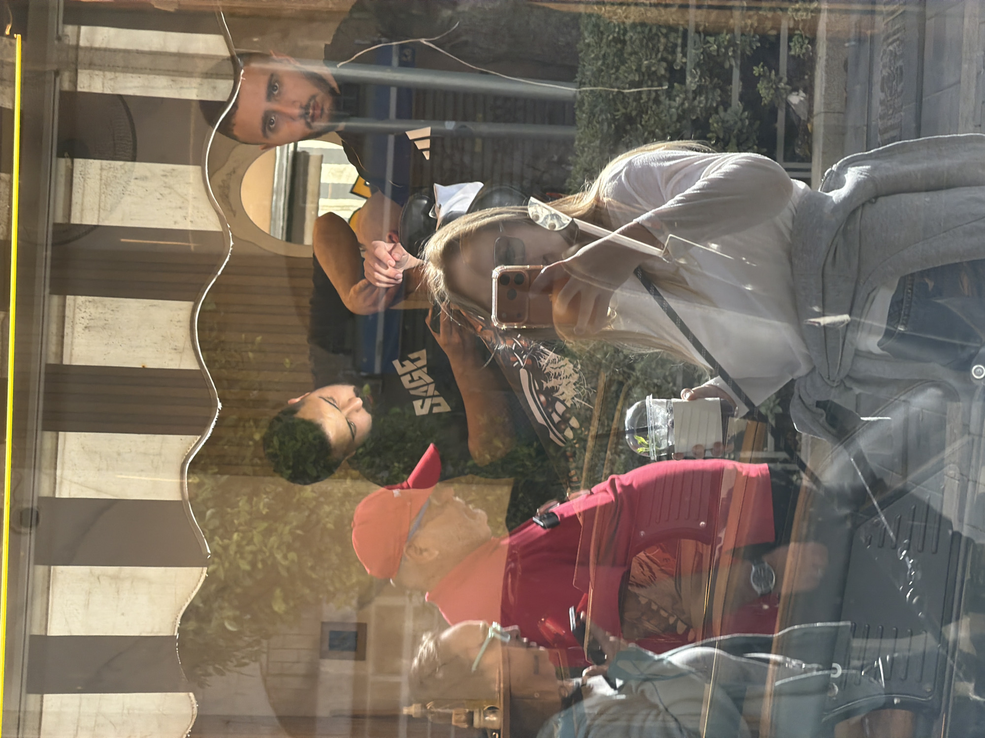 The image is a reflection in a window, likely a shop window or bus shelter. The reflection shows several people and parts of the environment.
People:
* A man with a beard looks directly at the "camera".
* A younger man is wearing a black shirt and looking down.
* A woman is holding a phone and appears to be taking a picture.
* There is another man wearing a red shirt and orange cap, and a woman wearing glasses.
Objects:
* A phone is being held.
* A plant in a clear container is visible.
Environment:
* Behind the window, trees and buildings are visible.
* The yellow side of a bus shelter.
Location Clues (Possible):
* Without more visual context, it's difficult to pinpoint the exact location in Tel-Aviv. Elements of the environment such as street furniture or architectural styles could provide further clues.