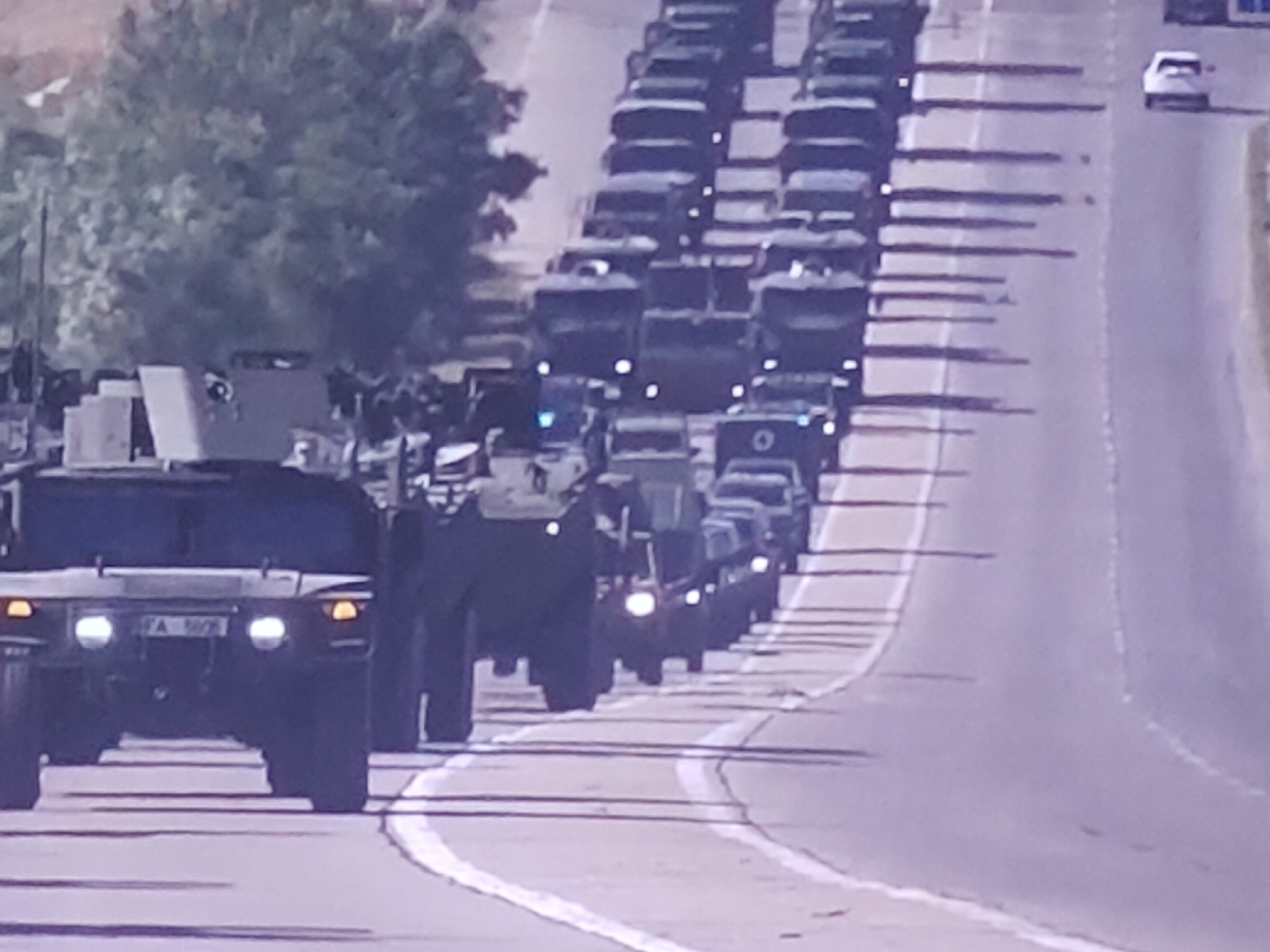 The image displays a procession of military vehicles on a paved road, likely in an urban or semi-urban setting, given the presence of what appears to be foliage in the background and a white car on the opposite side of the road. The primary focus is on a convoy of dark-colored military vehicles, including what appears to be a Humvee in the foreground, followed by several other trucks and armored vehicles. The vehicles are moving in the same direction, suggesting a convoy or movement of troops. The road has painted white lines, indicating lanes. The lighting suggests it is daytime, possibly late morning or afternoon, with distinct shadows cast by the vehicles. There is no clear indication of the specific location within Molenbeek-Saint-Jean - Sint-Jans-Molenbeek, Belgium, in the provided image. No individuals are clearly visible within or around the vehicles, and there are no overt signs of conflict or engagement. The scene appears to be a controlled movement of military hardware.