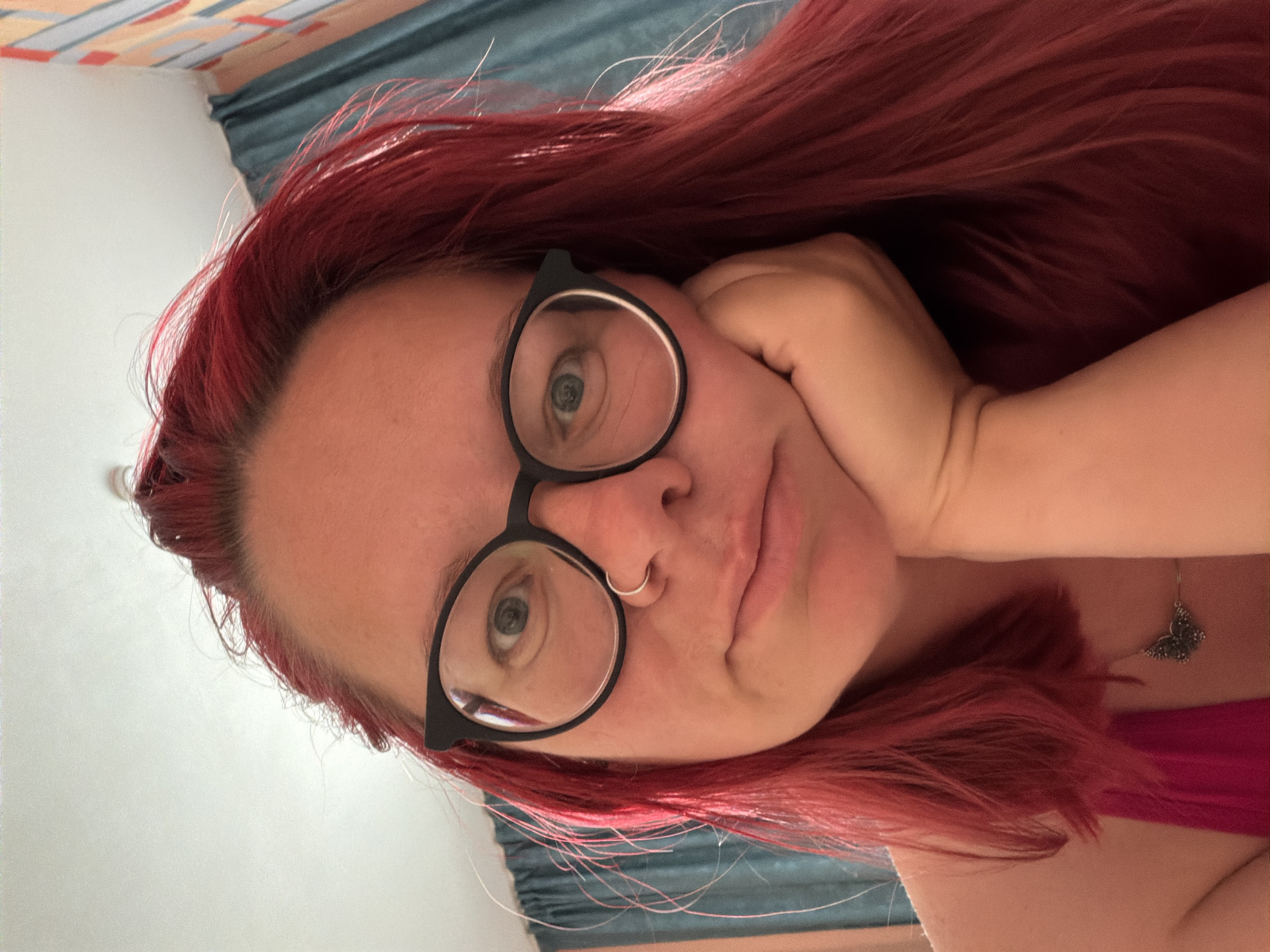 A portrait of a woman with red hair, wearing glasses and a nose ring, taken in an interior setting. She is resting her head on her hand and looking at the camera. A butterfly pendant is visible on her chest. The location is Chaikovsky, Russia. The background shows curtains and possibly a wall with patterns.