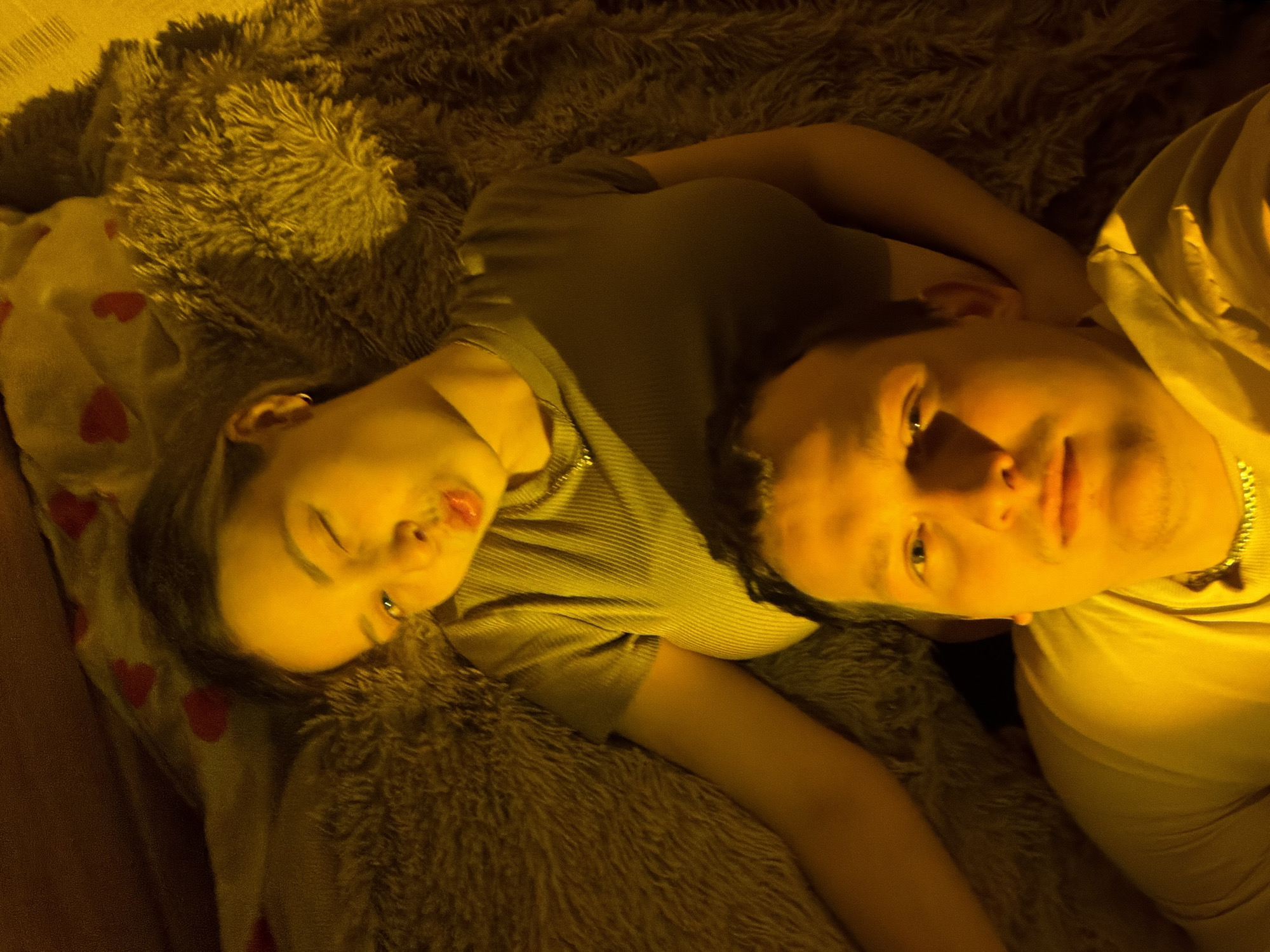 Two individuals, a female and a male, lie close to each other on a textured surface with a plush blanket, and a patterned pillow with red hearts. The location is not identifiable as being in the city of Chaikovsky, Russia. The female lies on her side with her eyes closed. She is wearing a collared shirt and a chain necklace. She has multiple piercings. The male is wearing a shirt and chain necklace. He lies with his head oriented towards the viewer and is looking into the camera. The scene appears to be dimly lit with an amber-colored light source.