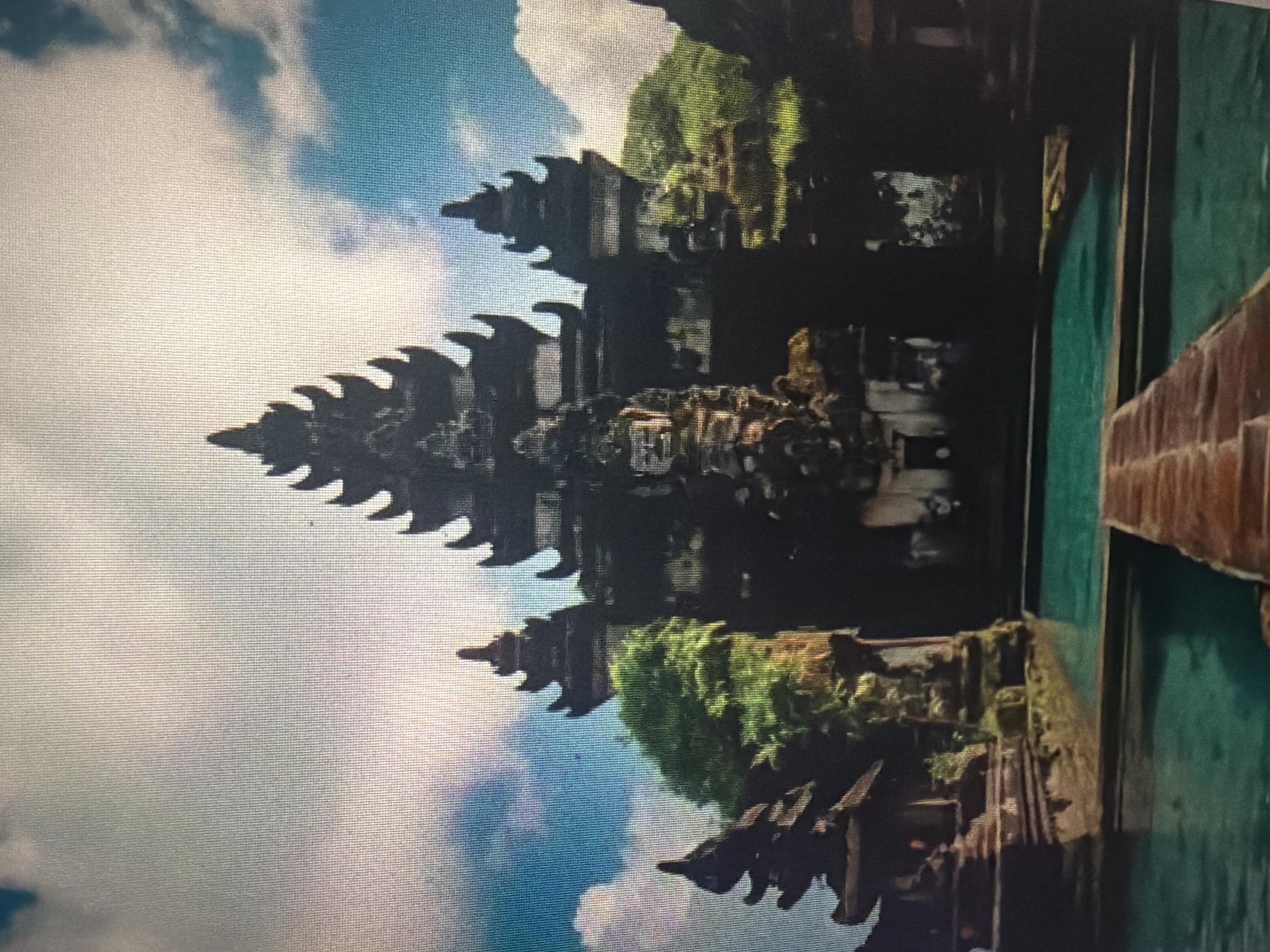The image shows a dramatic and ornate dark stone temple complex with tiered roofs, set against a bright, cloudy sky. A tranquil, turquoise pool of water is in the foreground, with a wooden walkway leading towards the temple. Lush green foliage surrounds the structure, suggesting a tropical or subtropical environment. The architecture is intricate and features decorative carvings, indicative of Southeast Asian temple design. The scene evokes a sense of grandeur and serenity. This location is not in Baan Mae Nam, Thailand.