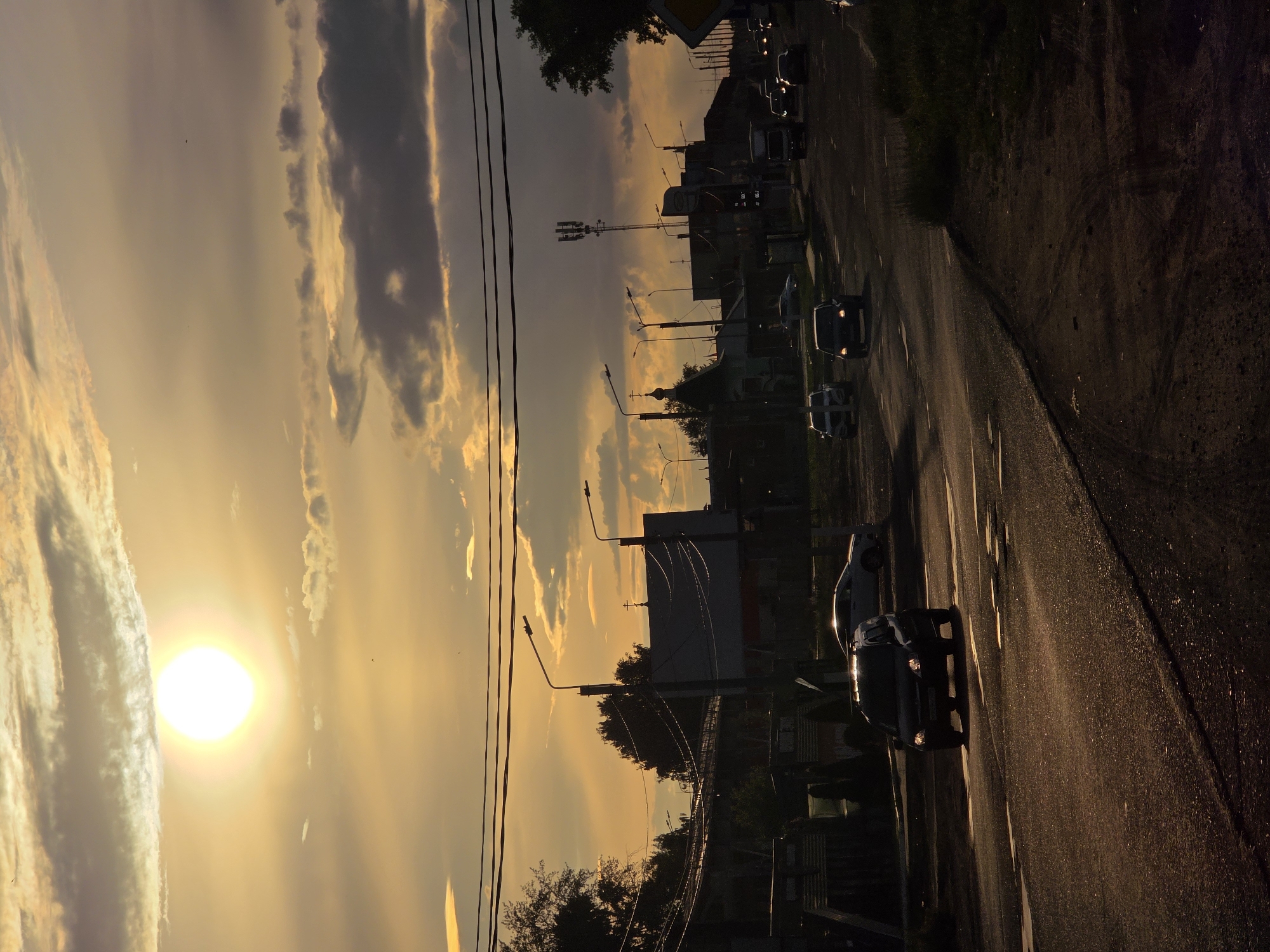 The image depicts a street scene at sunset in Chaikovsky, Russia. The sun, a bright orb, is low in the sky, casting a warm, golden light across the clouds and the wet asphalt road. Several cars are visible on the road, some driving and others parked. The scene includes utility poles with wires, streetlights, trees, and buildings lining the street, suggesting an urban or semi-urban environment. The overall mood is serene, amplified by the soft hues of the sunset and the quiet atmosphere. The road appears to have recently been rained on, as evidenced by the reflections of the car headlights on the wet surface. There is no visible text in the image.