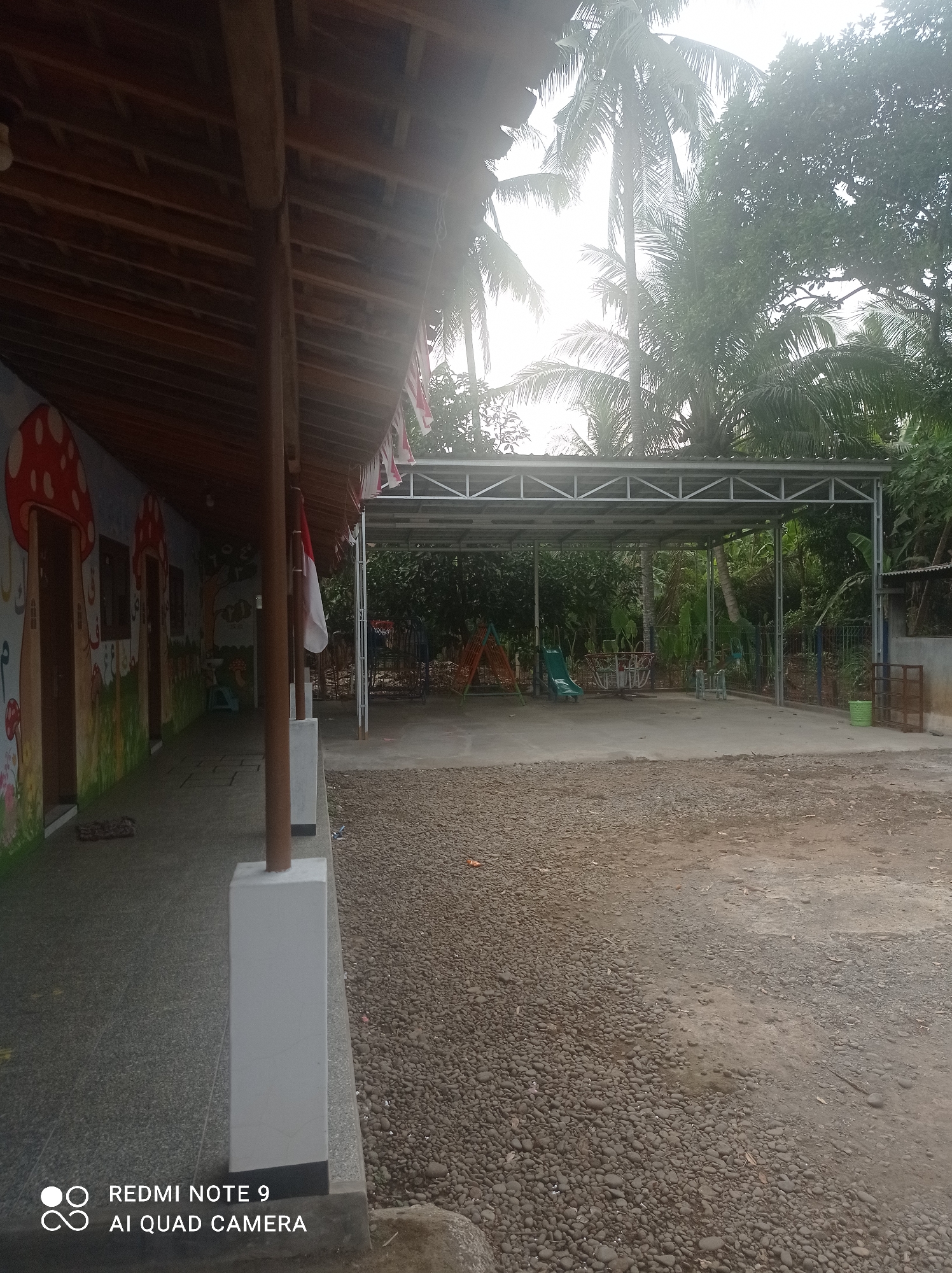 The image depicts an outdoor area, possibly a playground or schoolyard, in Cipawon, Indonesia. On the left side of the frame, a building with a wooden roof structure and brightly painted walls featuring mushroom and nature-themed murals is visible. A covered walkway runs along the side of the building. An Indonesian flag is prominently displayed, attached to a pole in front of the walkway.
The central and right portions of the image show an open concrete area covered by a metal canopy. This area contains playground equipment, including a green slide, an orange and red swing set, and a colorful circular ride. Beyond the covered area, a dense natural environment with trees and foliage is present, suggesting a tropical or sub-tropical location. The ground in front of the covered area is covered with small stones and gravel, with some damp patches indicating recent moisture. A green plastic bin and a metal fence are also visible on the right side. The overall impression is a functional outdoor space with elements of both constructed facilities and natural surroundings.