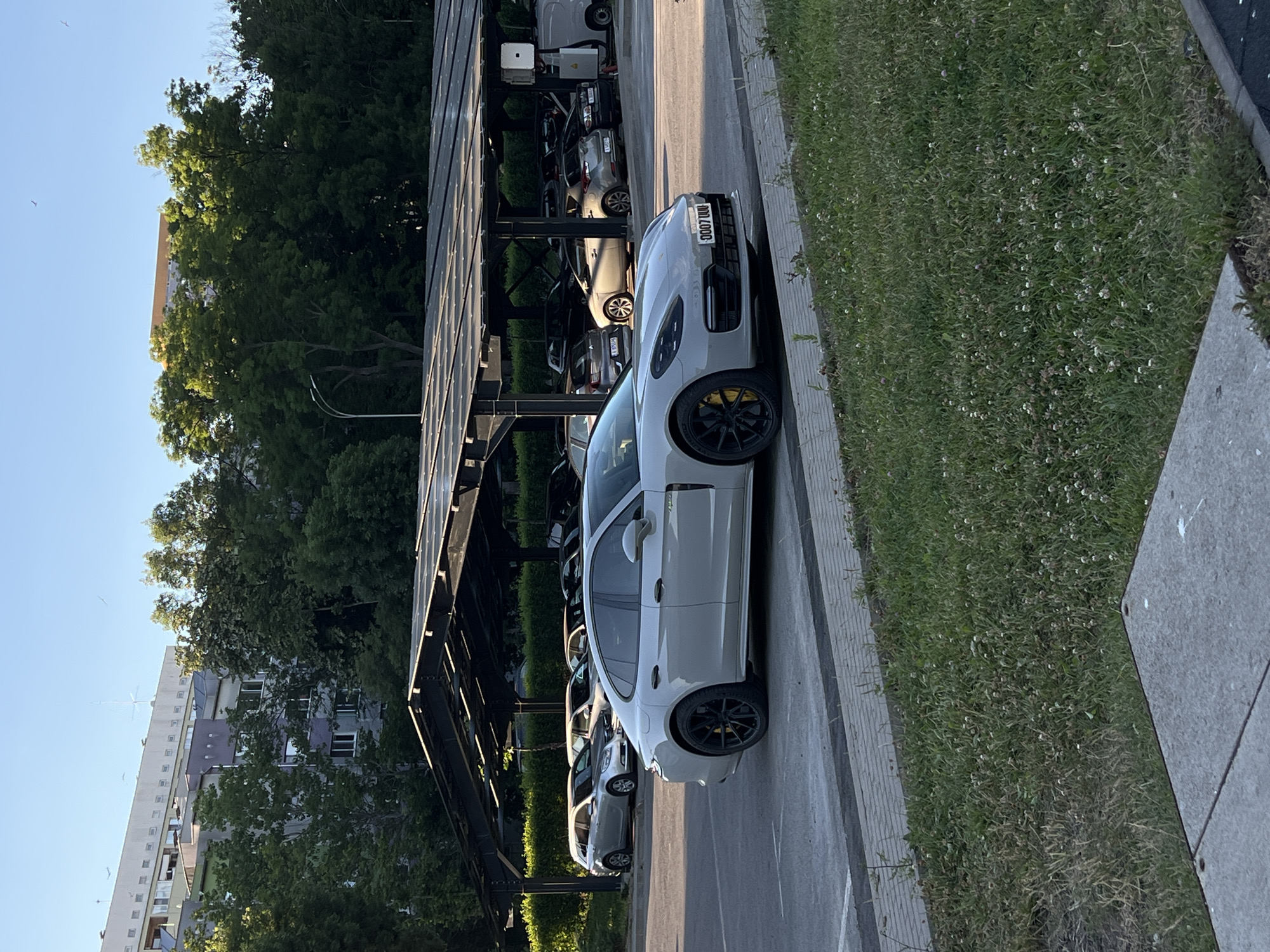 The image shows a light gray Porsche Panamera parked in a lot with a solar panel canopy over several other vehicles. The car has black wheels with yellow brake calipers. The license plate reads "0007 UU". In the background, there are trees and a building. The setting appears to be an urban environment, possibly in Burgas, Bulgaria, given the architecture and type of vehicles. The lot is paved, with a grassy area in the foreground. There are no people visible in the image. The lighting suggests it is either early morning or late afternoon.