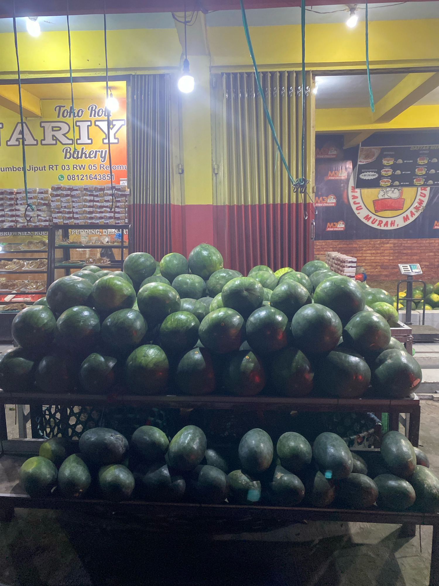 The image depicts a street-side fruit stand at night in the city of Kediri, Indonesia, specifically in the Rejowinangun area as indicated by the address "Jiput RT 03 RW 05 Rejomu" visible on the sign. The primary focus is a large display of watermelons, stacked on wooden shelves. The watermelons are predominantly dark green and appear to be ripe. Behind the watermelons, shelves containing baked goods are visible through a glass display, suggesting the stall also sells bakery items. A sign in the background advertises "Toko Roti ARIY Bakery," indicating the business name. The lighting in the scene is artificial, coming from overhead bulbs, casting a yellowish glow. There are no people clearly visible in the foreground or interacting with the fruit. The environment suggests a commercial area with storefronts and street lighting.
