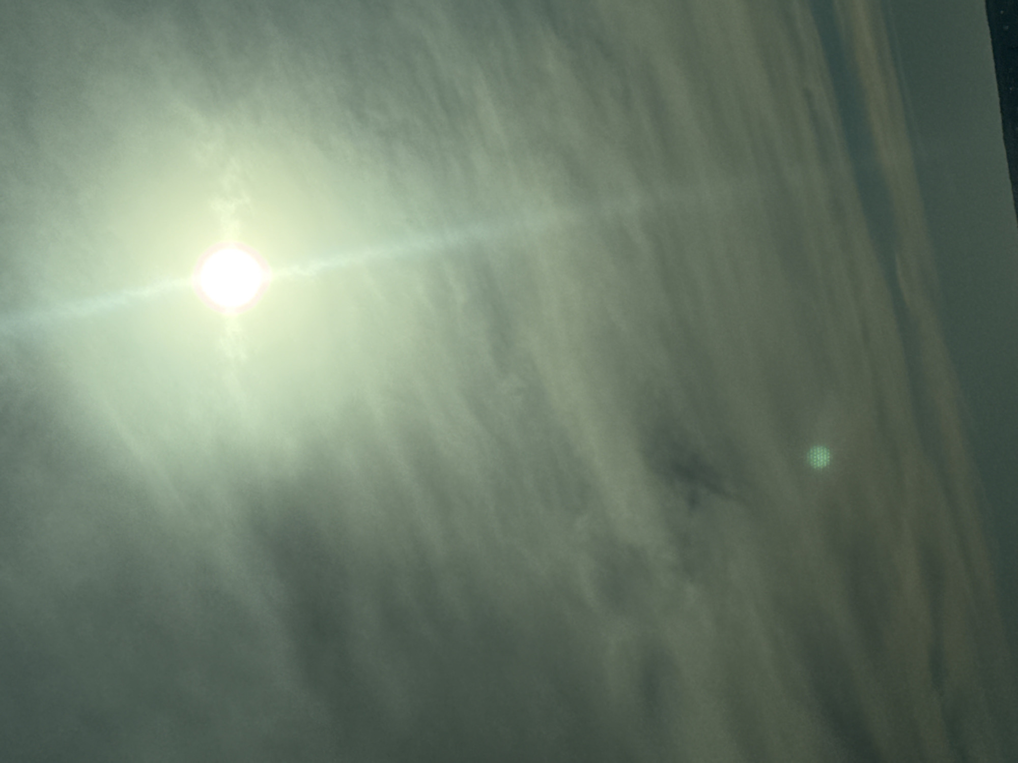 The image shows a view of the sky. A bright sun is visible through thin clouds, producing a strong glare. The clouds are streaky and textured. The lower part of the image is slightly darker than the upper part. There is a faint, slightly out-of-focus, circular green artifact in the lower right quadrant of the image. The location in the city of Пальники, Russia is unknown.