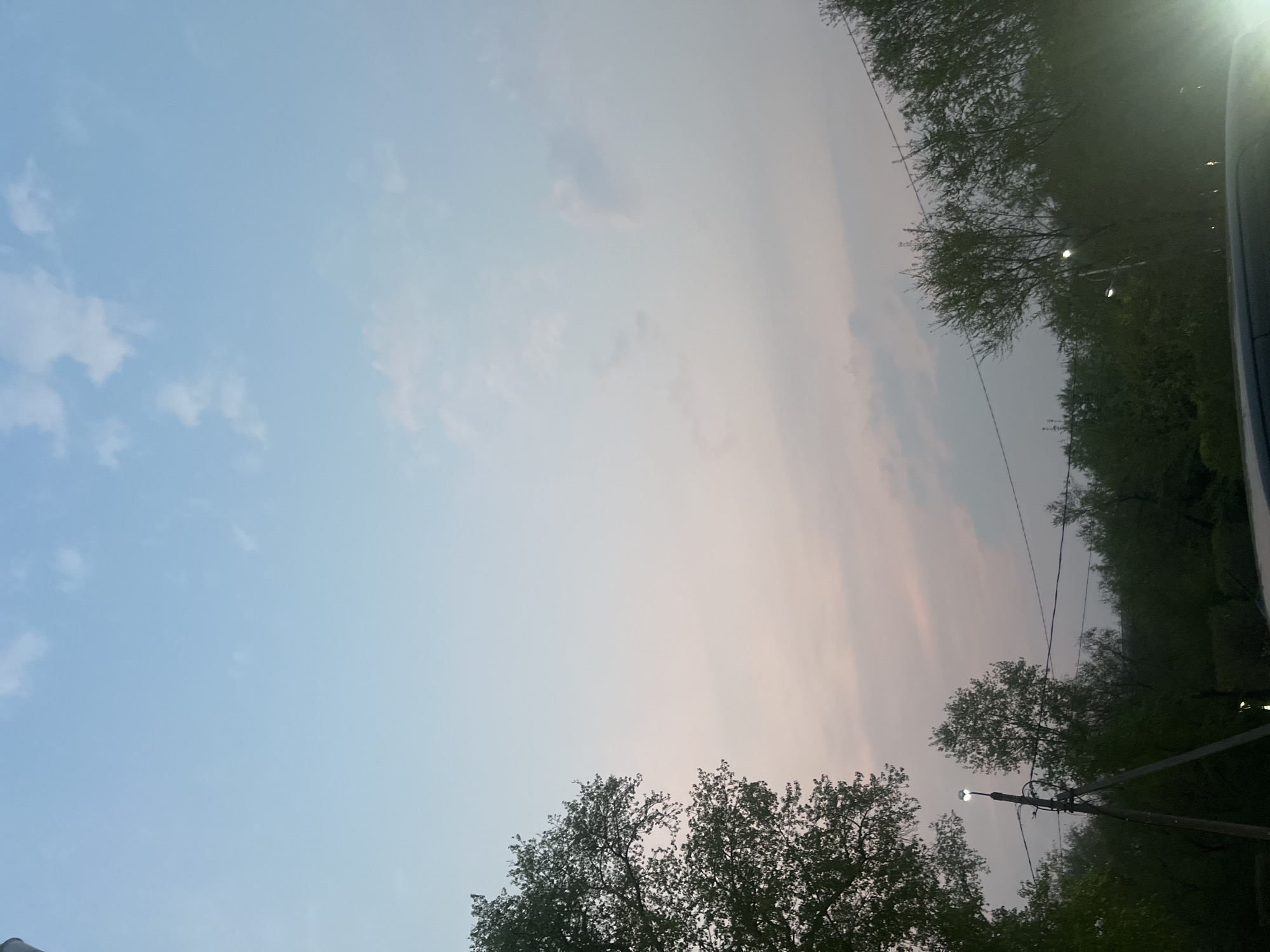 The image shows a portion of the sky and trees. The sky is predominantly blue with some wispy clouds. Near the horizon, the sky transitions to a pinkish hue, suggesting either sunrise or sunset. The trees are silhouetted against the sky, with leaves appearing dense and dark. A utility pole with a street light is visible, and the light is illuminated, indicating it is twilight or night. The perspective suggests the photo was taken from ground level, looking upwards. The location is Unknown, Unknown.