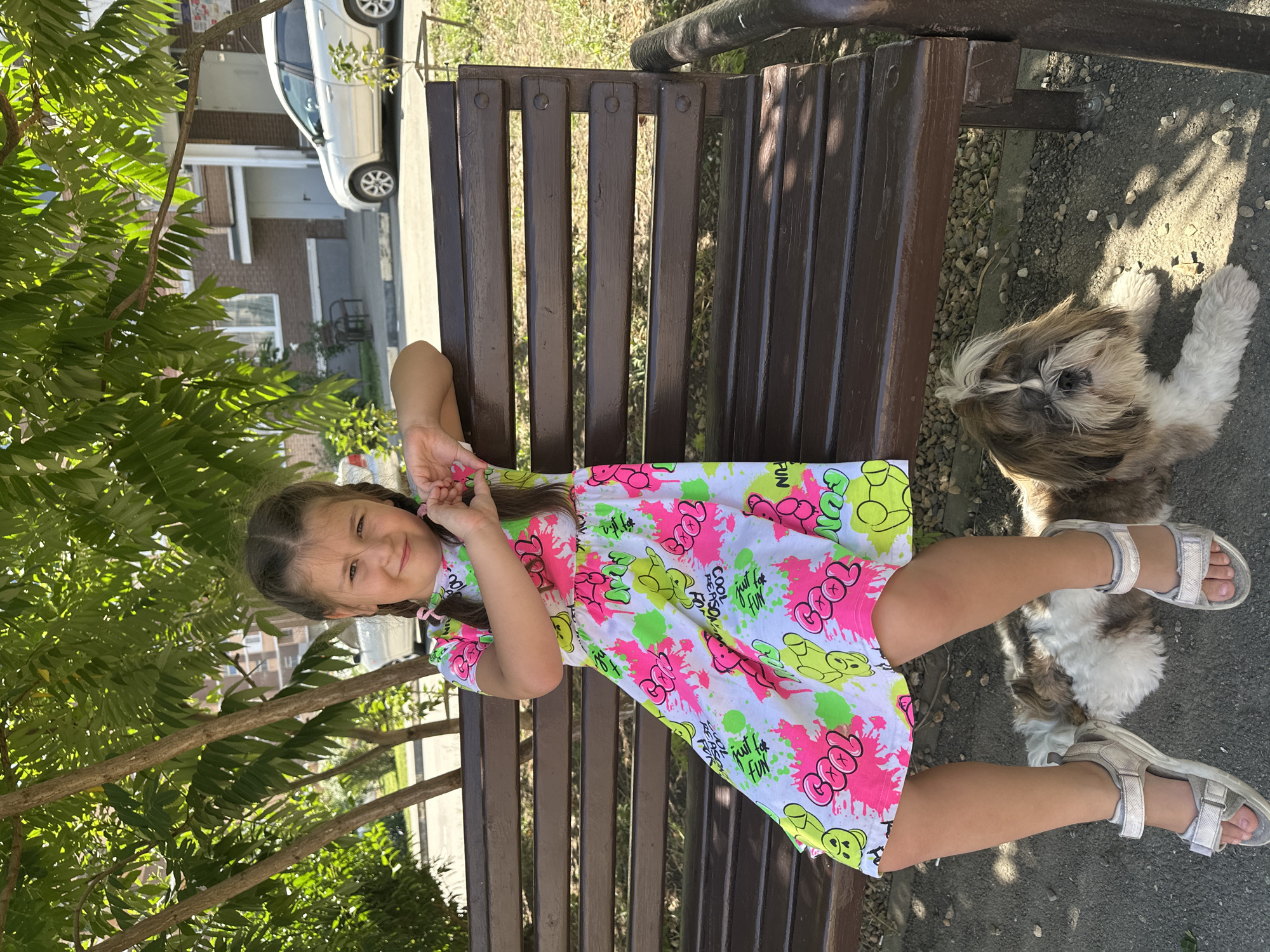 A young girl with brown hair in braids, wearing a white dress with pink, green, and yellow cartoon prints and silver sandals, sits on a dark brown wooden park bench. She is making a heart shape with her hands over her chest. To her right, lying on the ground, is a Shih Tzu dog with brown and white fur. The background shows a paved area with shadows from trees, some gravel, and a building with a car parked in front. The location appears to be outdoors in a park or residential area.