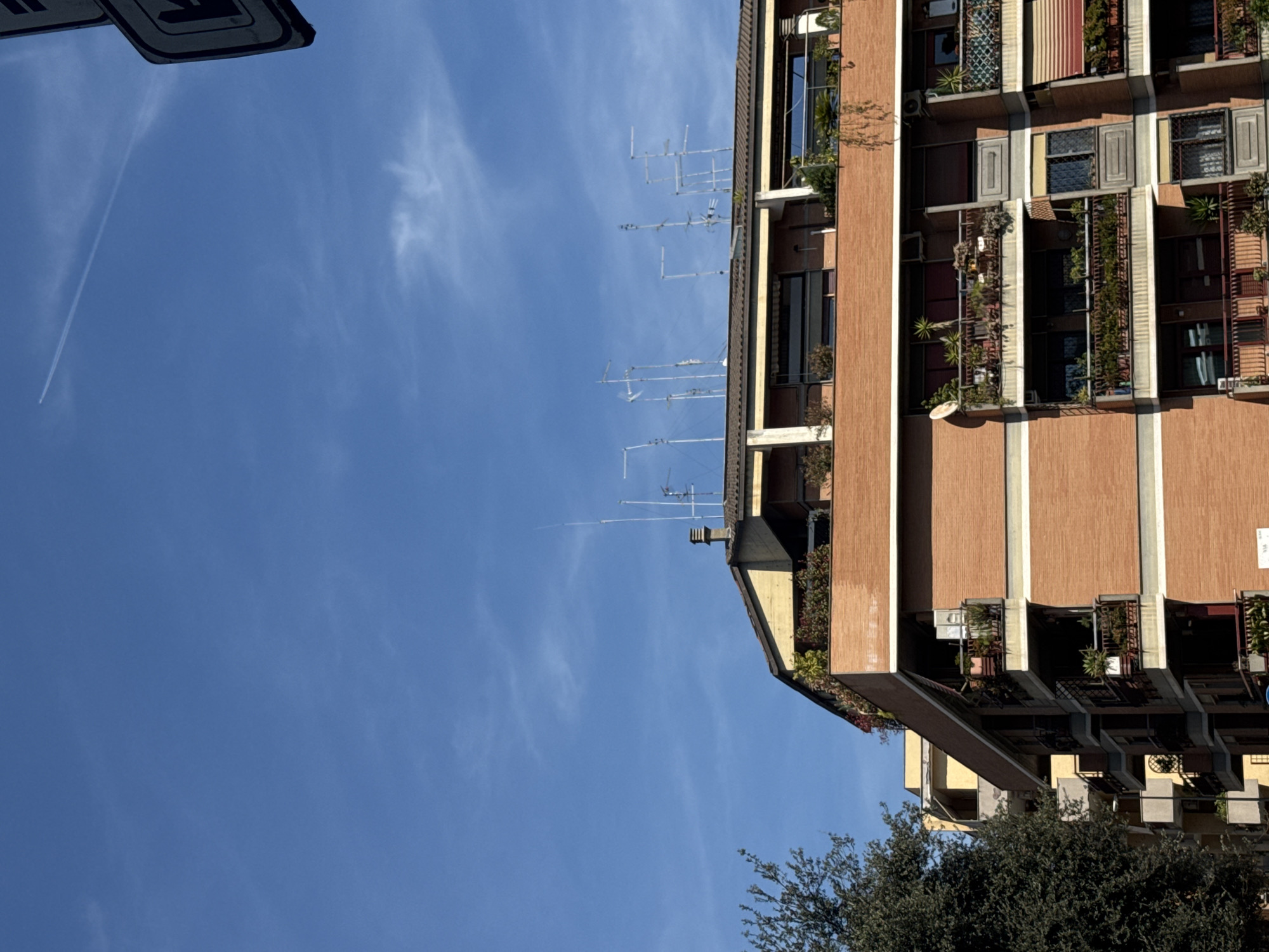 A low-angle shot of a building against a blue sky with some faint contrails in Rome, Italy. The building is multi-story with a brick facade and light-colored concrete accents. Many of the windows are lined with plants. There are numerous television antennas on the roof. At the bottom of the frame, a tree obscures part of the building. The scene is well-lit with a clear sky.