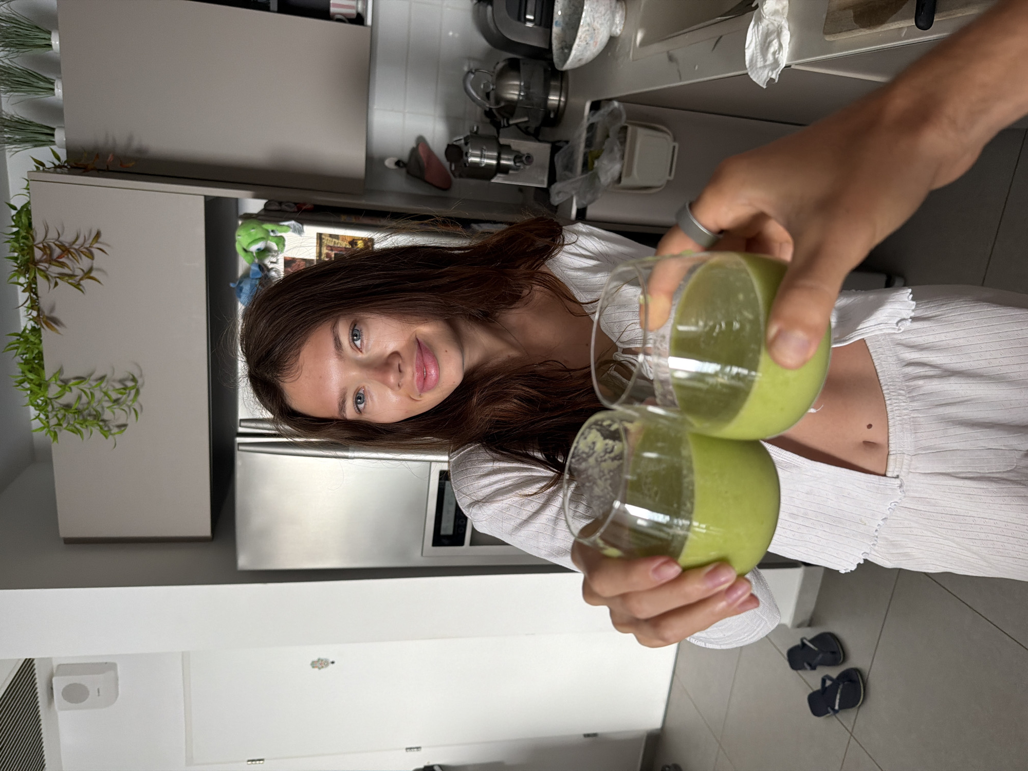 The image captures two people toasting with glasses filled with green smoothies, likely in a residential kitchen. One person, a woman with auburn hair, holds a glass up and looks toward the camera. She is wearing a white top. The other person's arm is visible holding another glass up to toast. The kitchen has white cabinets, a stainless steel refrigerator, and various kitchen appliances are on the counter. A pair of flip flops are on the tile floor. Although there are plants on the cabinet, the location is uncertain without other information. There is no forensic data visible in the image. Tel Aviv, Israel is not ascertainable from the image.
