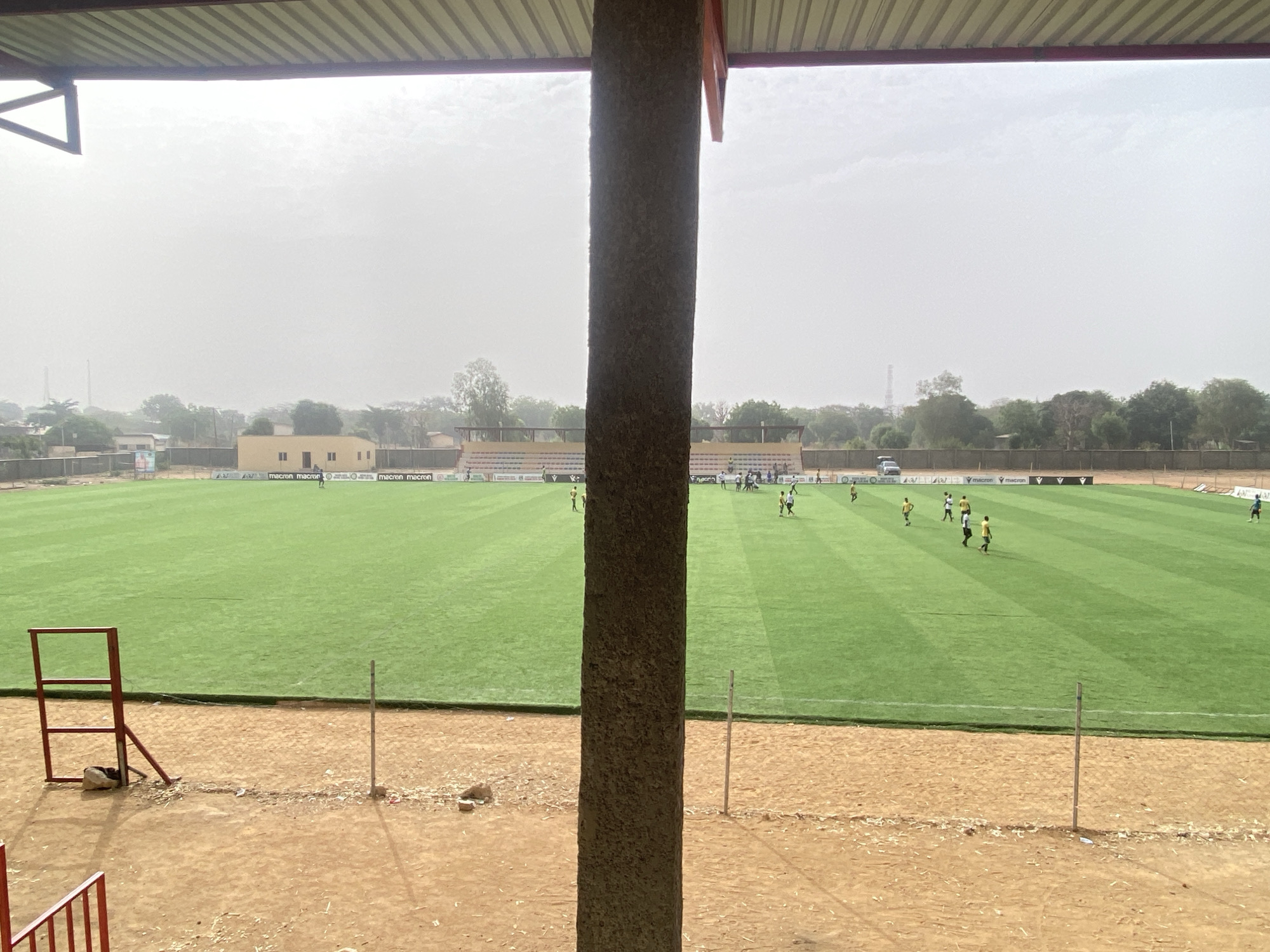 The image shows a soccer field in Tudun Wada, Nigeria, seen from under a covered structure with a support column in the foreground. The field is green and has players in yellow and white uniforms scattered across it. A white vehicle is parked behind the field near a building, and a fence lines the perimeter. In front of the field is a dirt area with some metal structures on the left. The sky is overcast.
