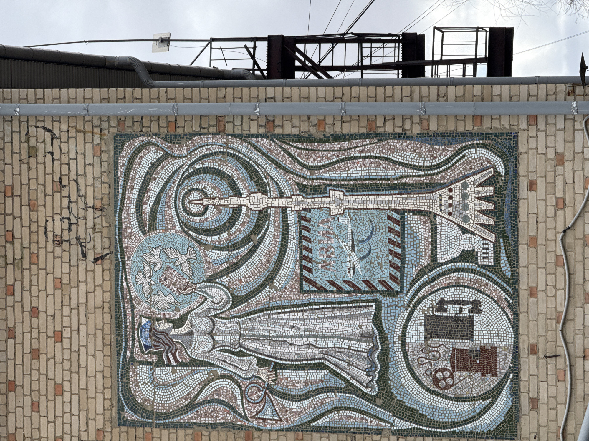 A mosaic mural is mounted on a brick wall. Above the wall are pipes and electrical equipment on the roof of a building. The mural depicts several symbolic figures. The top left has a circle of white doves. The mosaic continues with a reclining figure beneath the doves. Above the figure is a long, horizontal instrument pointed toward the upper right corner. Above the instrument is a square with striped corners. In the bottom right is a round circle containing a depiction of some kind of machinery. The mosaic is on the side of a building in Borzya, Russia.