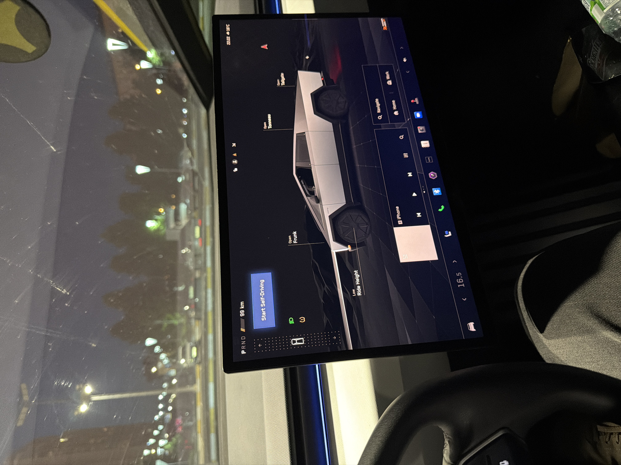 The image, taken in Moscow, Russia, captures the interior of a vehicle at night, specifically focusing on a large central touchscreen display. Part of a person's leg, clad in grey pants, is visible in the bottom right, along with a section of the steering wheel, indicating a driver's perspective.
The main subject is the vehicle's infotainment screen, which displays a detailed 3D rendering of a Tesla Cybertruck. The screen shows various control options for the vehicle, including buttons to "Open Frunk," "Open Tonneau," and "Open Tailgate." A "Start Self-Driving" button is prominent, alongside indicators for "PRND" and "99 km" (likely remaining range). The "Ride Height" is set to "Low."
Other information displayed on the screen includes the current time, "22:22," and the outside temperature, "16°C," confirming it is nighttime. Icons for navigation ("Q Navigate," "Home," "Work") and media playback ("iPhone," play/pause, skip) are also present, with the volume indicated as "16.5."
Through the vehicle's window, the dark urban environment is visible, illuminated by numerous streetlights. Reflections of these lights and smudges are present on the glass. A bottle and possibly a bag can be partially seen near the screen on the right.
