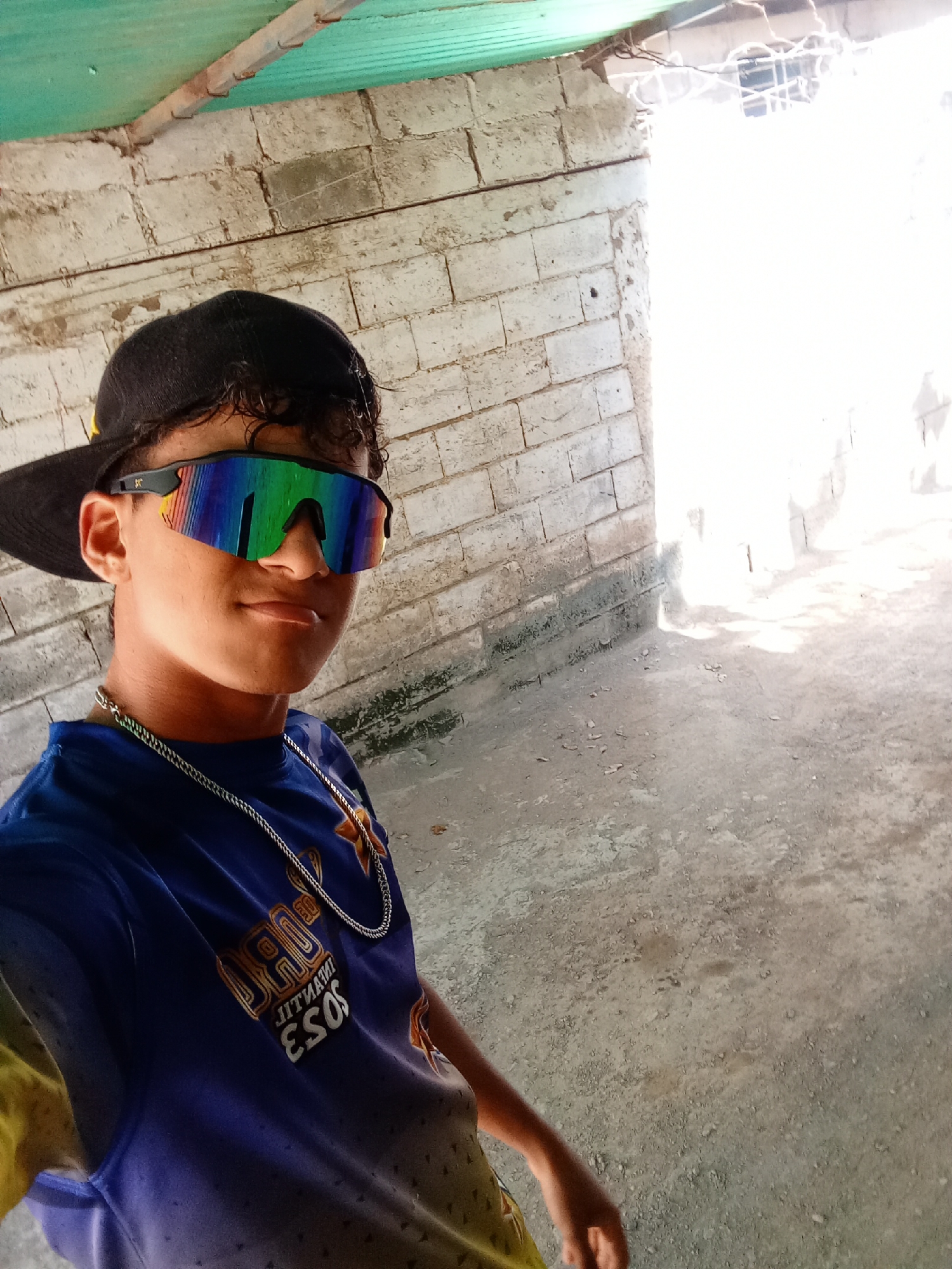 A young male wears a backward black baseball cap, reflective sunglasses, and a dark blue and yellow t-shirt with a silver chain around his neck. The background features a white brick wall, a green corrugated metal roof, and a concrete floor. The location is likely in or around the city of Juan Griego, Venezuela, as indicated in the prompt. The scene appears to be an outdoor, covered area.