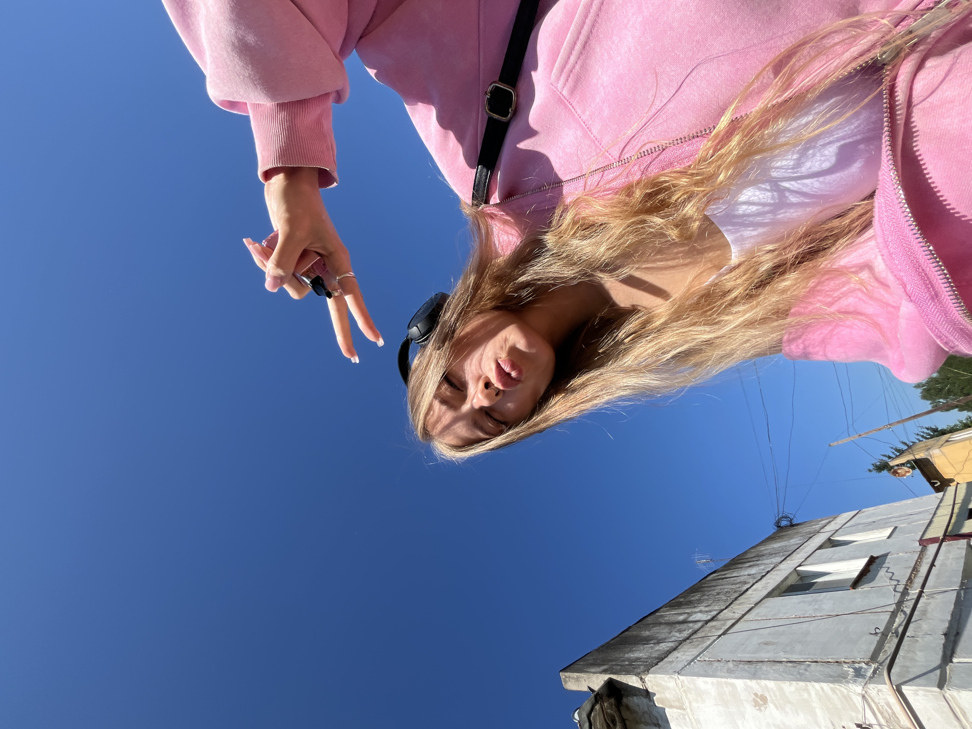 A young woman with long blonde hair, wearing headphones and a pink zip-up hoodie over a white tank top, pouts her lips and makes a peace sign with her fingers under a clear blue sky. She is in the city of Foky, Russia. To the left of the woman, a weathered, multi-story building with concrete walls and visible signs of aging stands against the sky. The building has several windows and some debris on its exterior. In the background, telephone wires are strung across the scene, and a few trees are visible to the right. The image is taken from a low angle, looking up at the woman and the sky.