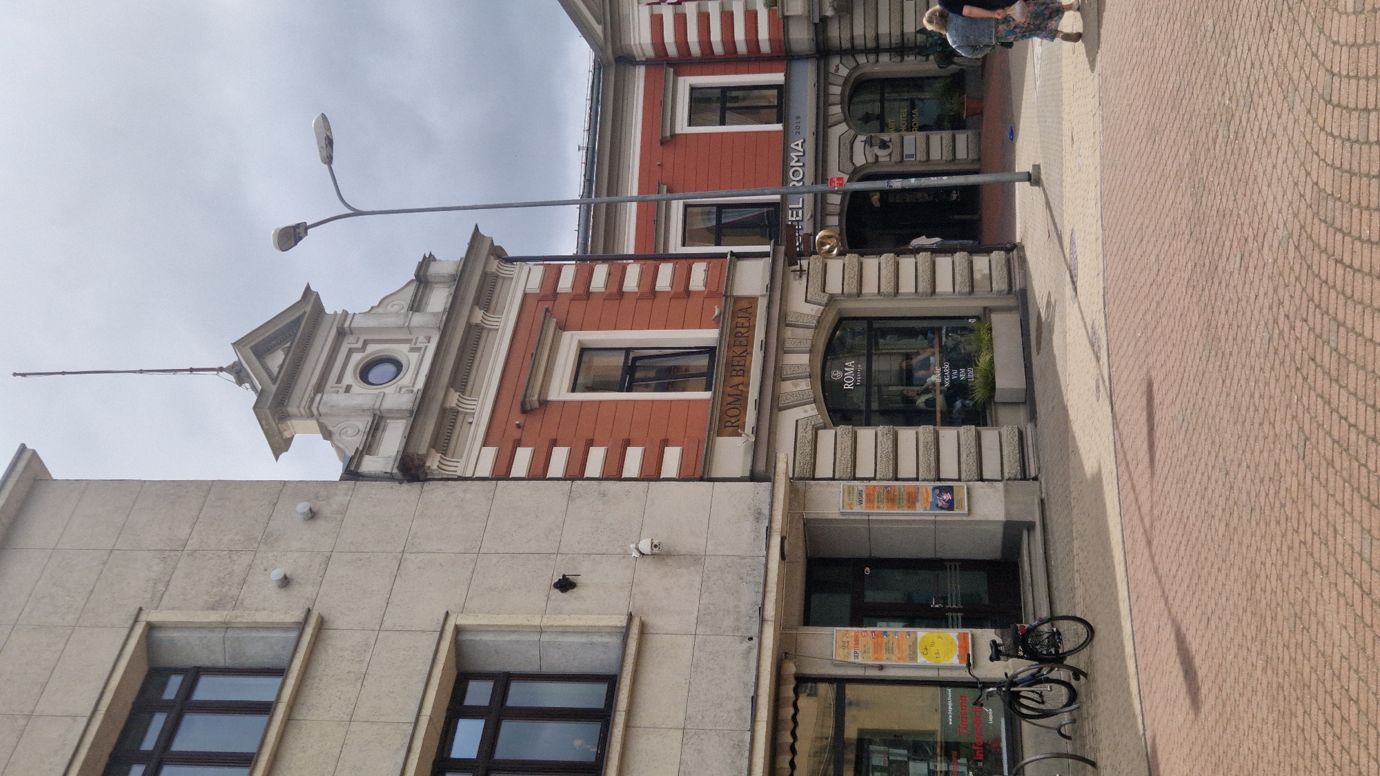 The image shows the exterior of a building in the city of Liepaja, Latvia. The building features a red facade with white decorative elements and a prominent sign that reads "ROMA BEKEREJA". Below this, at street level, is a storefront with the "ROMA" logo visible. To the right of "ROMA BEKEREJA", another sign indicates "HOTEL ROMA 2019".
In front of the building, a paved sidewalk with a brick pattern is visible. A black bicycle is parked on the left side of the sidewalk. Two streetlights are present, one tall and slender, the other shorter and with a broader lamp.
Two individuals are walking away from the building on the right side of the frame. The person in front is wearing a patterned dress and a light-colored jacket. The person behind is wearing a denim jacket and darker pants.
The sky is overcast, suggesting a cloudy day. The architecture suggests an older building with some modern elements, likely in a commercial or mixed-use area of the city. Posters are visible on the wall of the building to the left, one of which appears to advertise events for September.