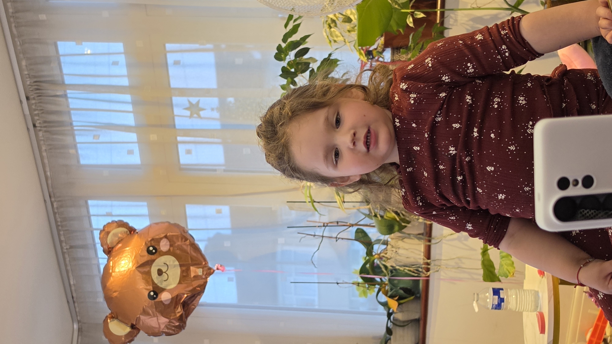 A tilted indoor scene shows a young girl holding a cell phone in front of her, possibly taking a selfie. She is wearing a long-sleeved burgundy dress with a white floral print. Beside her is a shiny, metallic balloon shaped like a brown bear. In the background, there's a large window with sheer curtains that reveals a view of buildings. There are also several potted green plants visible in the frame. The setting appears to be a domestic interior, with various household objects scattered around, including a water bottle. The location is likely a home interior, and there is no indication that this is London, United Kingdom.