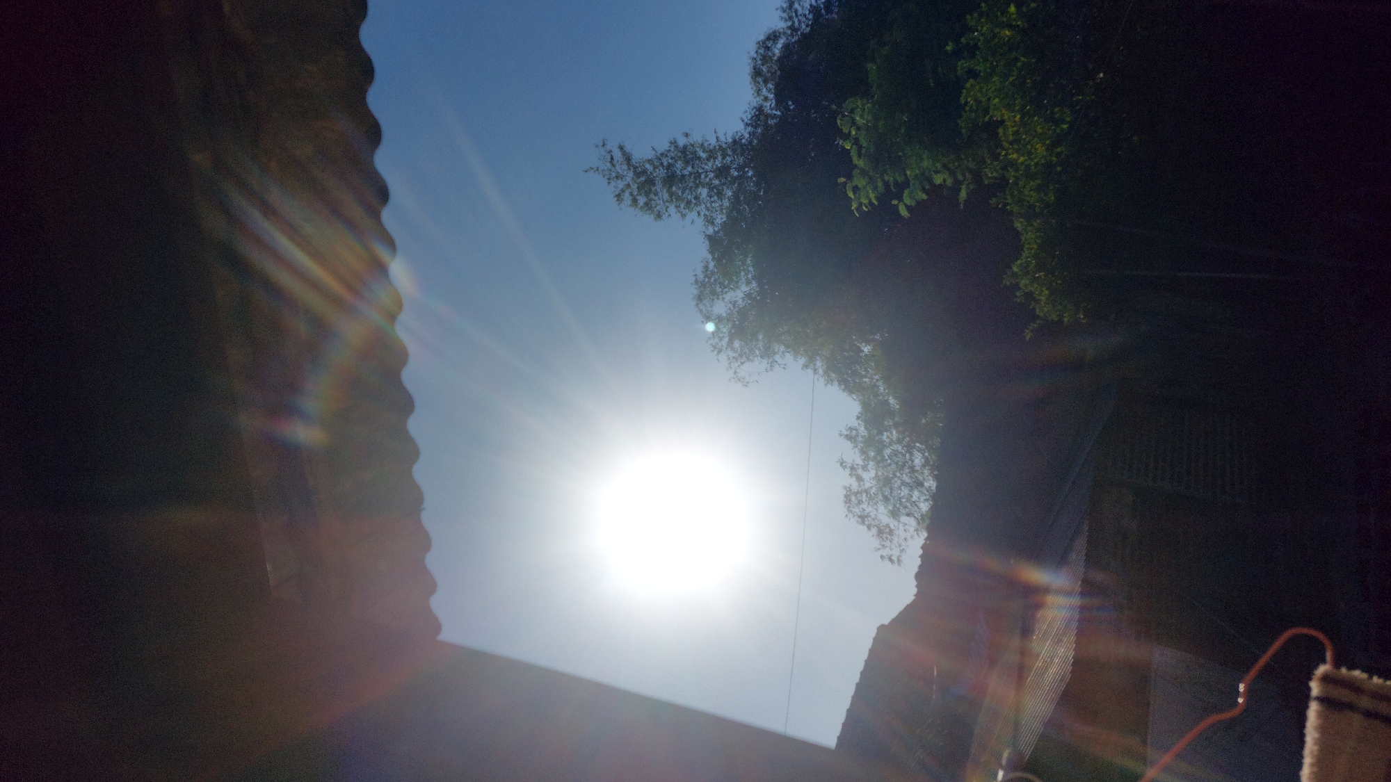 The image depicts a bright sun shining in a clear blue sky, creating lens flare and light streaks across the scene. In the foreground, a dark, corrugated roofline is visible, casting a shadow. Below the roof, the silhouettes of buildings and trees are discernible, with a hint of brickwork on one structure. The overall impression is of an outdoor environment, possibly residential, bathed in strong sunlight. The location is Jatirogo, Indonesia, and the scene is captured from a low-angle perspective, looking upwards towards the sky.