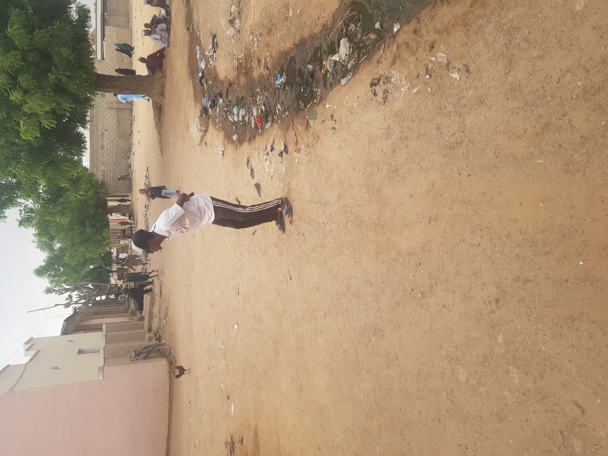 A medium, tilted, outdoor shot shows a street scene in Dutsinma B, Nigeria, with a man in the center doing push-ups on a dirt road. The man wears a white shirt, black sweatpants with white stripes, and black sandals. He faces away from the viewer towards other people further down the street.
To the left of the man, a tree stands next to a pink building with a bicycle parked outside. Further up the road, people sit and stand near a large tree and a brick wall. To the right of the man, a narrow, muddy ditch contains refuse. The ground is primarily dirt. The sky is overcast.
