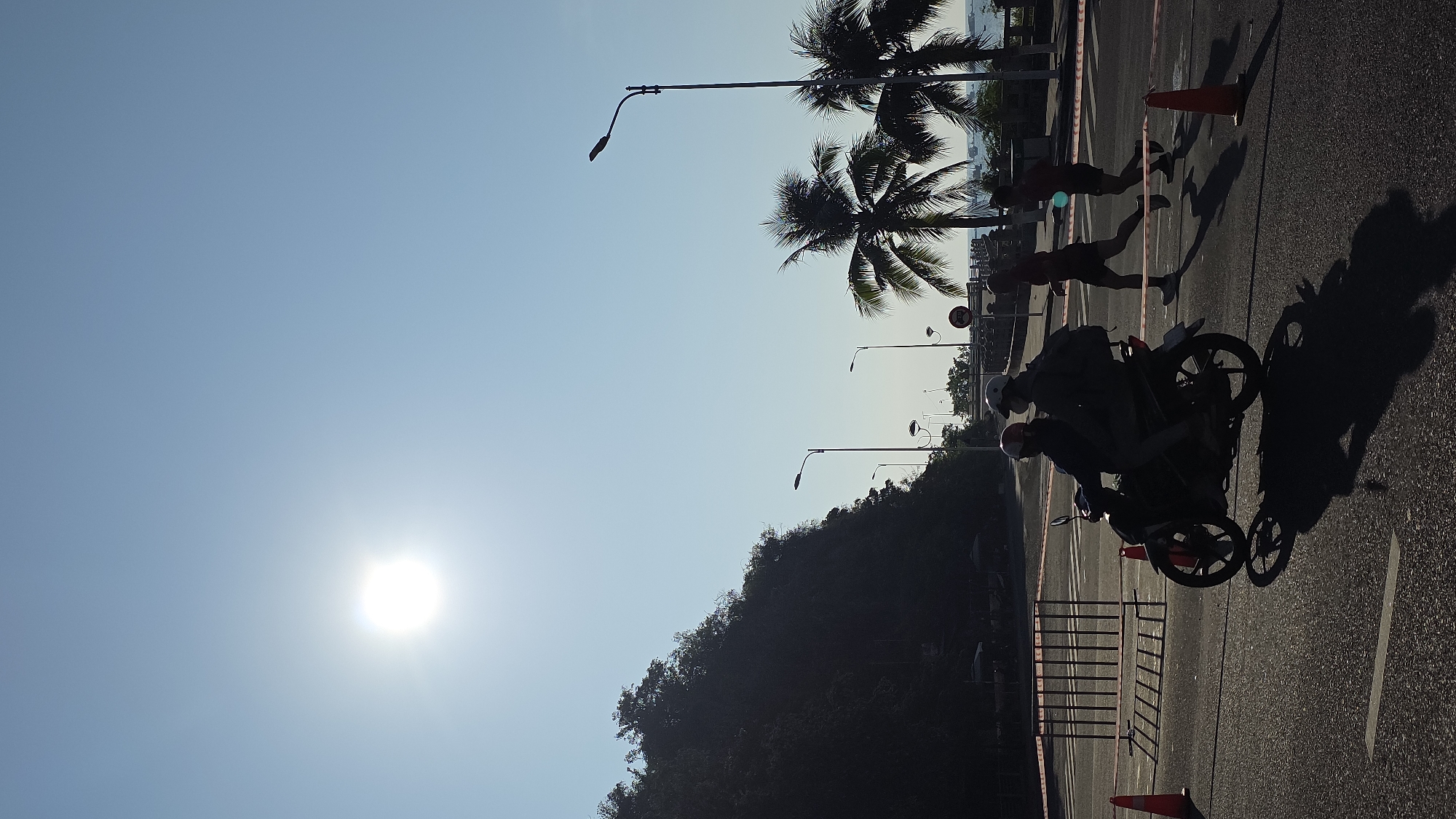The image shows a bright, sunny day, likely in the early morning or late afternoon due to the low angle of the sun in the sky. The setting appears to be a coastal road, identifiable by the presence of palm trees and a glimpse of the sea in the background. This is possibly Nha Trang, Vietnam.
Silhouetted figures are present on the road. Several appear to be running, while two individuals are riding a motorbike, also in silhouette. Traffic cones and a metal barricade suggest a restricted or controlled area, perhaps for a race or event. Streetlights line the road.
A densely vegetated hill rises on one side of the road, further suggesting a coastal setting. The sky is clear, and the dominant feature is the bright sun, which affects the visibility of the subjects.