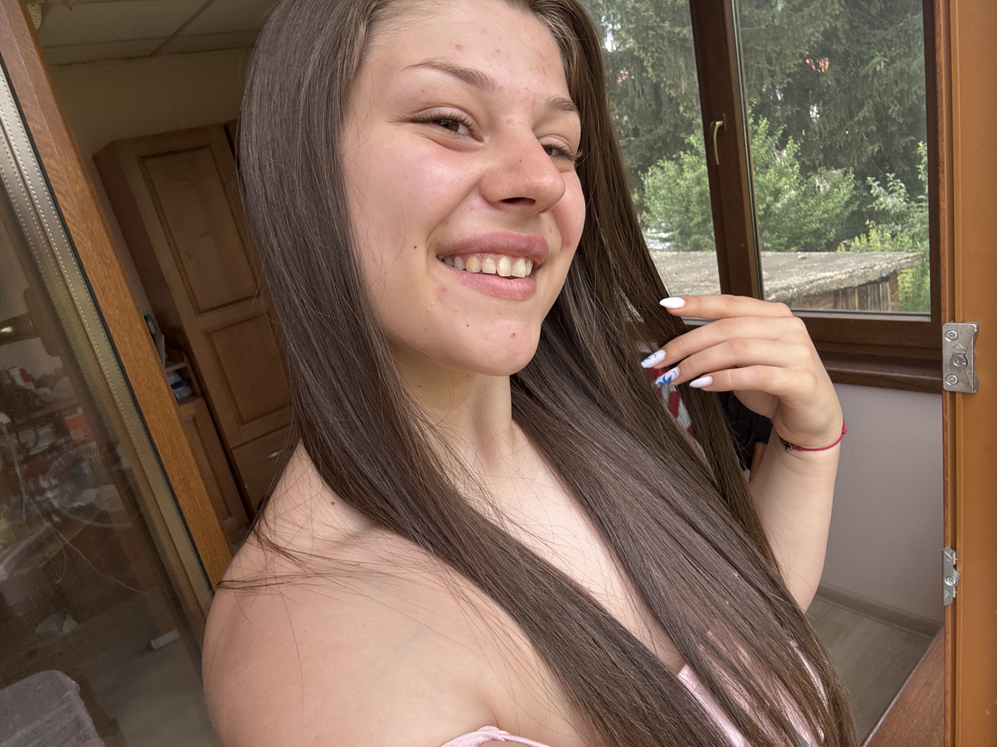 A young woman with long, dark brown hair is captured in a close-up, smiling self-portrait. Her gaze is directed slightly away from the camera, and her mouth is open in a wide, joyful smile, revealing her teeth. Her skin appears to have some minor blemishes. She is wearing a light pink top, visible around her shoulders. Her right hand is raised, with white nail polish on her fingernails and a red bracelet on her wrist. The background is softly blurred, but a wooden framed window is visible behind her, showing green trees and foliage outside. To the left of the woman, a wooden cabinet with drawers is partially visible, suggesting an indoor setting. The lighting suggests it could be daytime, possibly late afternoon, given the soft natural light. The overall mood is cheerful and relaxed. No visible text is present in the image.