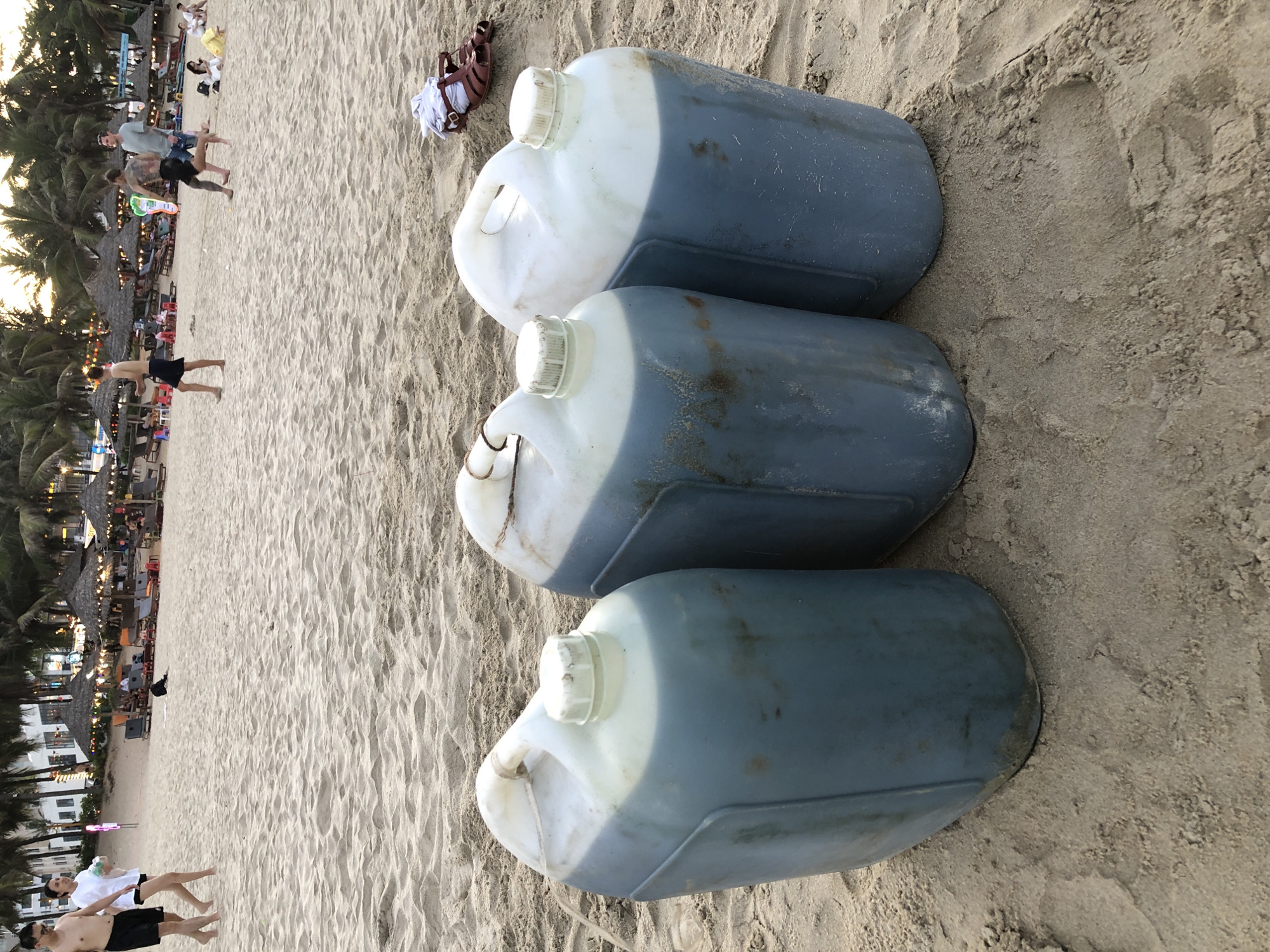 The image depicts a sandy beach, likely in Đà Nẵng, Vietnam, with several people in the background. In the foreground, three large, blue plastic jerrycans filled with a dark liquid are placed on the sand. One jerrycan is on the left, and two are to its right, positioned slightly in front. The jerrycans have white spouts and handles, with some appearing to have a white plastic cap secured. The beach is dotted with palm trees and umbrellas, suggesting a resort or public beach area. People are visible in the background, some walking, some sitting under umbrellas, and others in groups. One couple is walking towards the left in the mid-ground. Further back, a few individuals are seen near the water's edge and around beach chairs. The sand shows footprints and patterns from the tide or wind. A pair of brown sandals and a white t-shirt are also present on the sand near the jerrycans on the right.