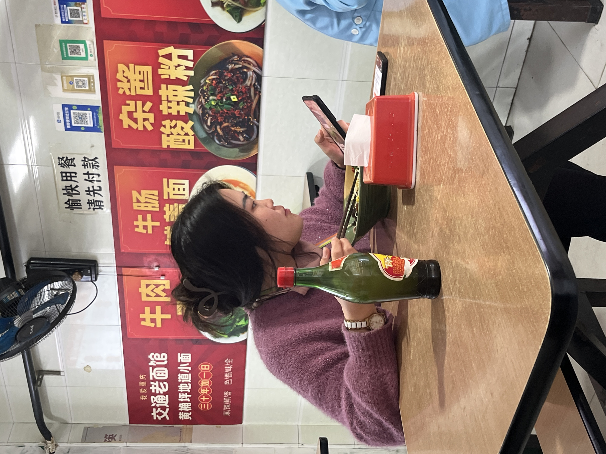 A sideways shot features a young woman sitting at a table in what appears to be a restaurant in Jiulongpo District, China. The woman, with dark hair pulled back in a clip, is dressed in a pink sweater and wears a watch on her left wrist. She is holding a smartphone and appears to be looking at it. A bottle of green liquid, possibly a condiment or sauce, is placed on the table next to her arm. Also on the table is a red tissue box and chopsticks in a wrapper.
Behind her, a wall display advertises various noodle dishes, including beef noodles, beef intestine noodles, and spicy noodles, in Chinese characters. QR codes are also visible on the wall, likely for mobile payment or ordering purposes. A small fan is mounted on the wall to the left.
