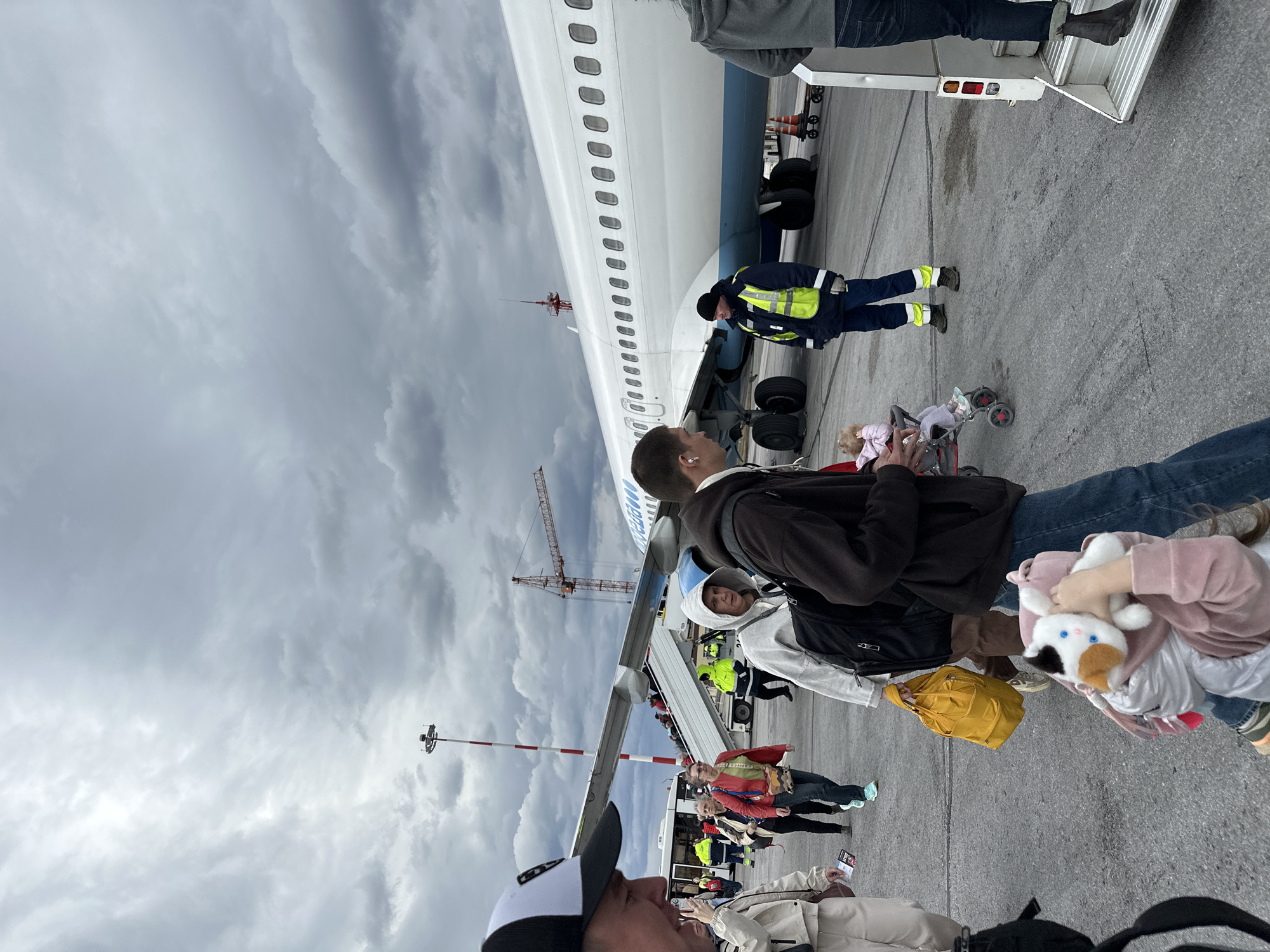 A daytime scene unfolds on an airport tarmac, showing numerous passengers queuing to board a large white and blue passenger aircraft under a heavily overcast, cloudy sky.
In the foreground, several individuals are partially visible, including one person wearing a black and white baseball cap. Further back, a line of passengers waits to ascend the mobile passenger stairs leading into the aircraft. Among them, a person in a dark jacket carries a yellow bag, and another holds a white and brown stuffed animal. A baby stroller containing a doll is also present on the tarmac.
The airplane dominates the midground, its white fuselage featuring rows of windows and a blue underside. The partial text "UTair" is visible on its side near the front. Airport ground staff, dressed in high-visibility yellow and blue jackets and dark pants, are positioned near the aircraft's landing gear and the base of the boarding stairs, overseeing operations.
The tarmac is dark grey asphalt. In the background, a large red and white construction crane and a tall light pole with aviation markings are visible against the gloomy sky. The weather appears cool and cloudy, characteristic of an overcast day. The overall activity depicted is the routine process of boarding an aircraft. The scene is located in Bolshoye Savino, Russia.