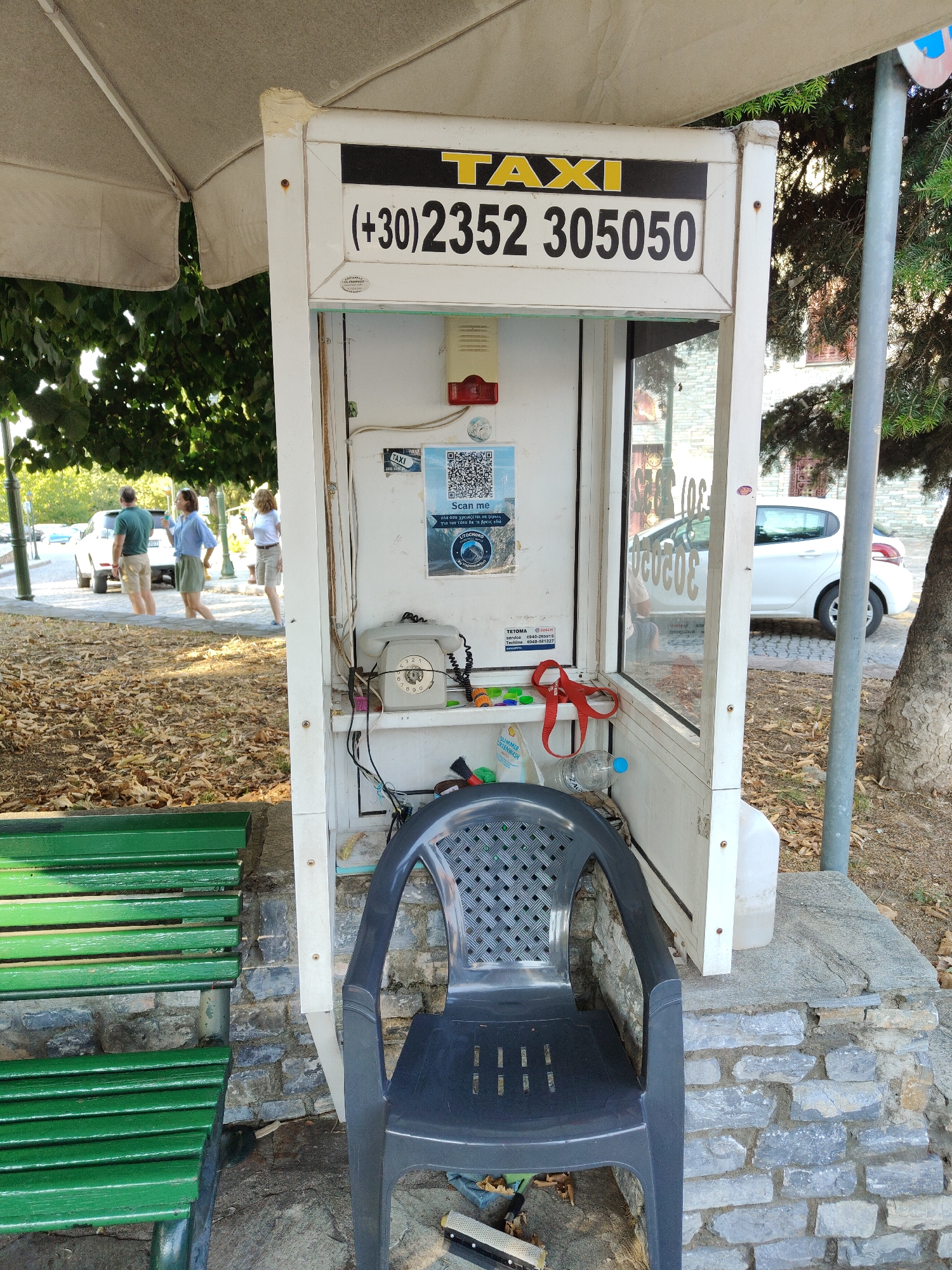 An outdoor scene in Litochoro, Greece, features a white, open-air taxi phone booth with a visible phone and a red plastic object resembling scissors. Above the phone booth, a sign displays "TAXI" in yellow letters against a black background, followed by the phone number "+30 2352 305050". Inside the booth, a rotary dial telephone is mounted on the wall. A QR code is also affixed to the wall. A dark grey plastic chair sits directly in front of the booth, and a green wooden bench is positioned to the left. In the background, three individuals stand near a white car parked on a paved area. The ground is covered with fallen leaves, and trees are visible to the right and behind the booth.