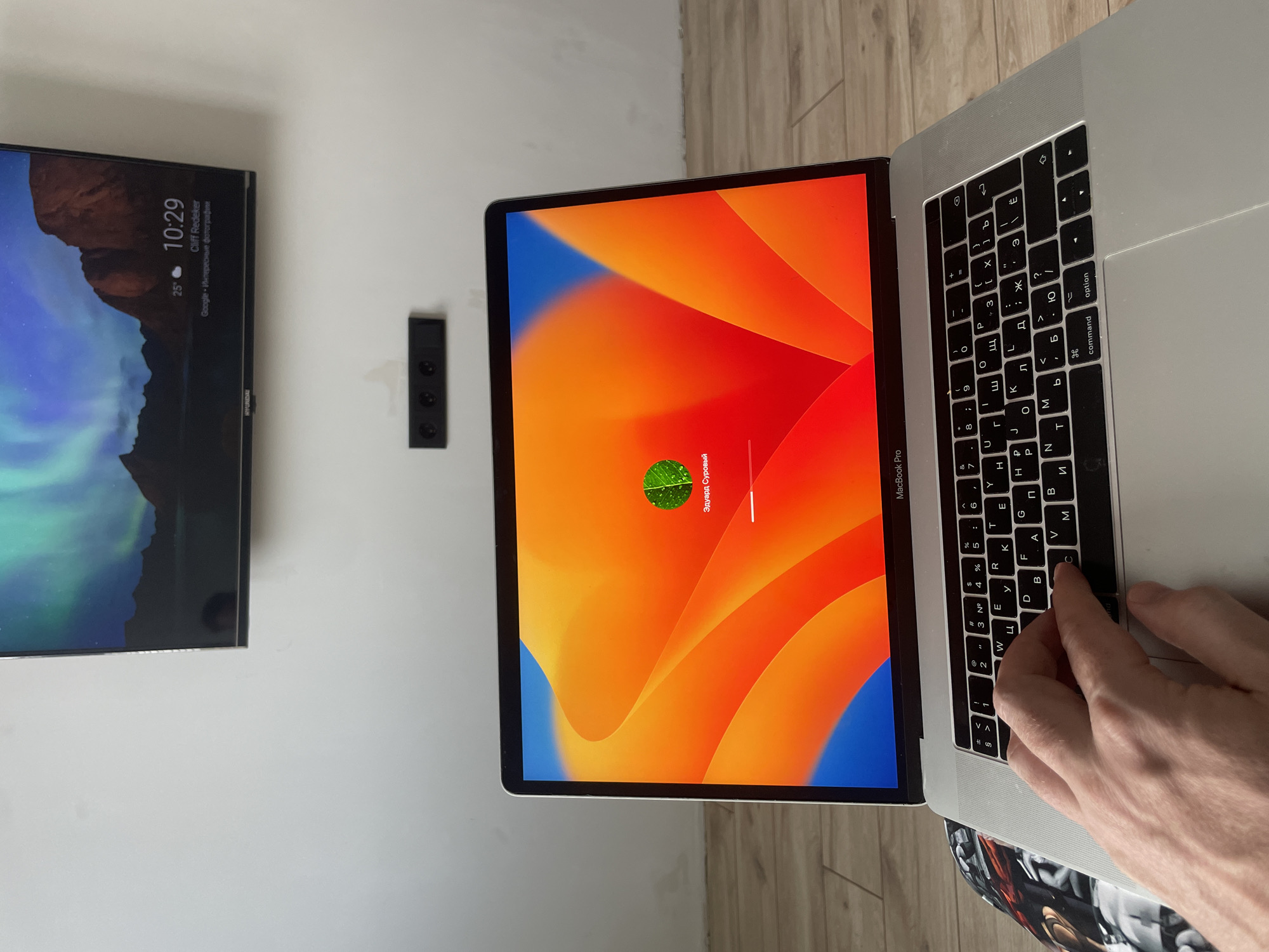 The image shows a laptop with a bright orange and blue abstract wallpaper. On the laptop screen, there is a circular green icon with a leaf design and text below it that reads "Эдуард Суровый". A progress bar is visible below the text, indicating loading. A person's hand is on the keyboard, with fingers positioned over the 'C' and 'V' keys. The laptop is a MacBook Pro. In the background, a television screen displays a colorful image of the aurora borealis over mountains, with the time "10:29" and temperature "25°" displayed. A black wall-mounted power outlet with four sockets is visible on the wall. The floor appears to be made of wooden planks. The scene is indoors, likely a room in a home or office. There is no indication of the city of Kazan, Russia, in the image.