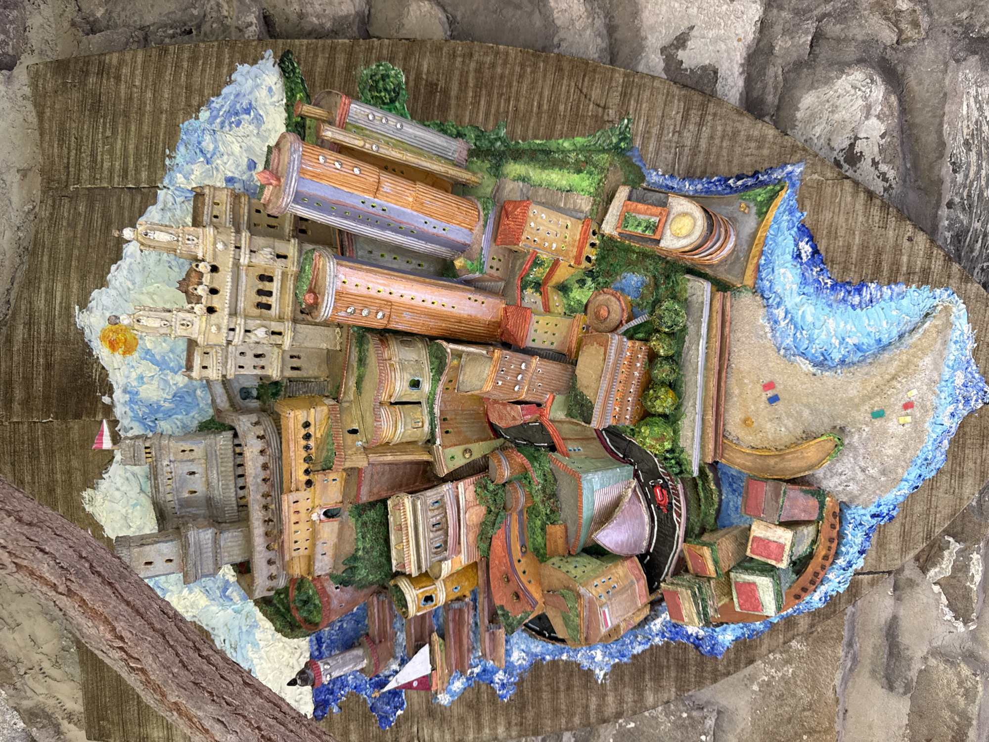 The image is a detailed, three-dimensional diorama depicting a stylized coastal city, possibly inspired by Dolceacqua, Italy, or a similar Mediterranean town. The diorama is mounted on a rustic, wooden board that resembles aged planks, framed by a rough, bark-like material on the left and stone on the right.
The city itself is a vibrant, colorful assemblage of buildings, structures, and natural elements, all rendered with thick, textured paint that gives the impression of a miniature world. In the upper left portion of the diorama, a large, grey fortress-like structure with turrets and battlements dominates the scene. A small, white sailboat with a red stripe is visible in the blue water to the left of the fortress. Adjacent to the fortress, a lighthouse stands tall.
Further into the city, a grand, palatial building with twin towers and arched windows is depicted. A bright yellow sun with radiating rays is painted in the sky behind this structure. Various other buildings of different shapes and sizes, some cylindrical and tall with visible window details, are clustered together. The architecture suggests a blend of historical and more modern elements. Buildings are painted in warm earthy tones, oranges, yellows, and muted reds, with green foliage interspersed throughout. Roads, painted in black and red, wind through the city, and a red sports car is shown on one of these roads.
The diorama's base appears to be a rough, wavy representation of the coastline, with blue paint simulating water. A sandy area with small, colorful flags is also present near the water's edge. The overall impression is of a bustling, picturesque coastal town, presented in a highly artistic and somewhat whimsical style. There are no visible people or discernible actions occurring within the diorama. The focus is entirely on the architectural and environmental representation of the miniature city.