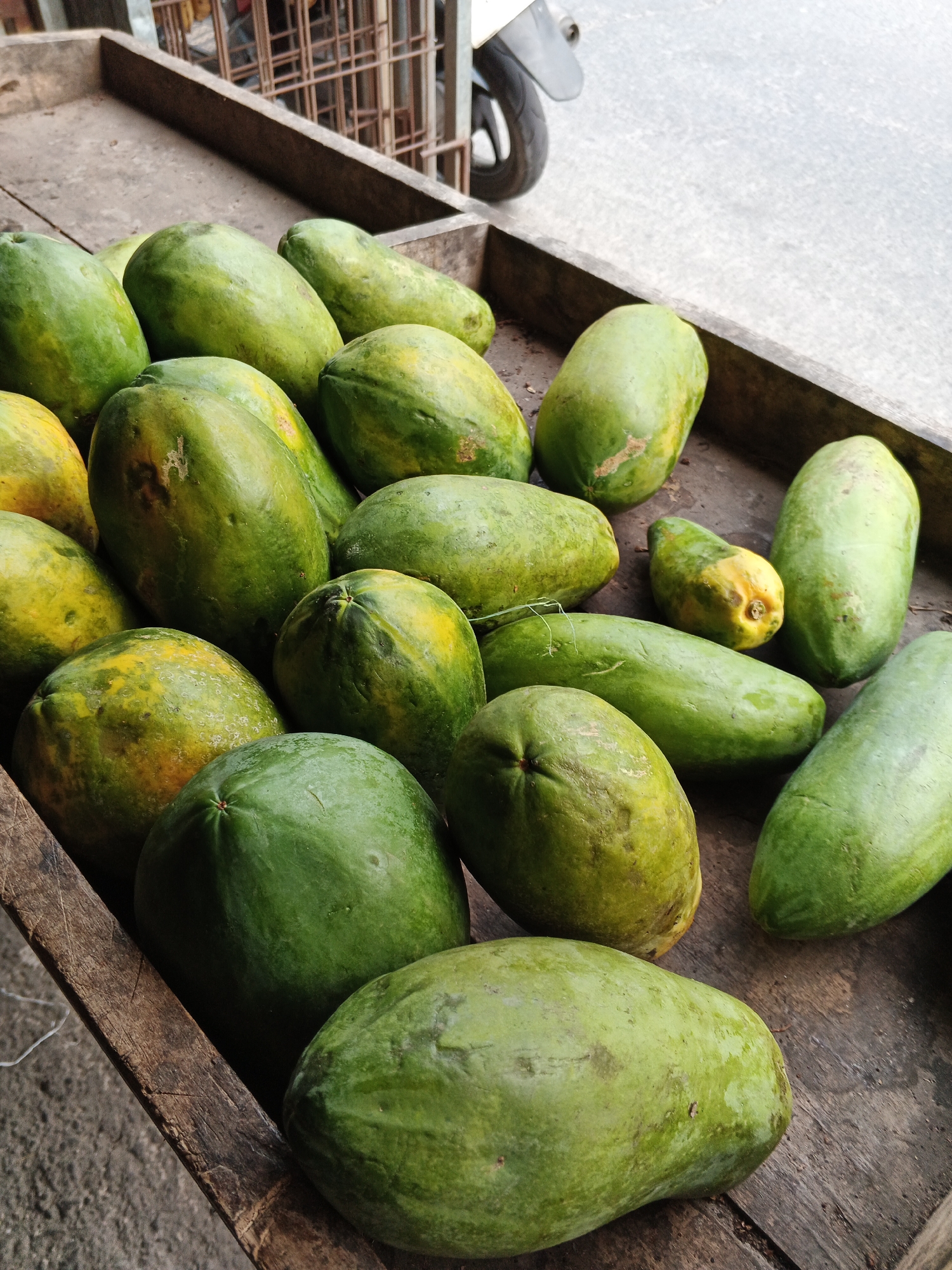 A close-up, high-angle shot shows a collection of green papayas, some with yellowing patches, resting in a weathered wooden crate. The crate is positioned on a textured surface, possibly concrete, with a blurred background revealing a metal grate structure and the rear wheel of a scooter. The papayas vary in size and shape, with some exhibiting slight imperfections on their skins. The lighting suggests an outdoor setting during the day. The scene appears to be in Gandasari, Indonesia.