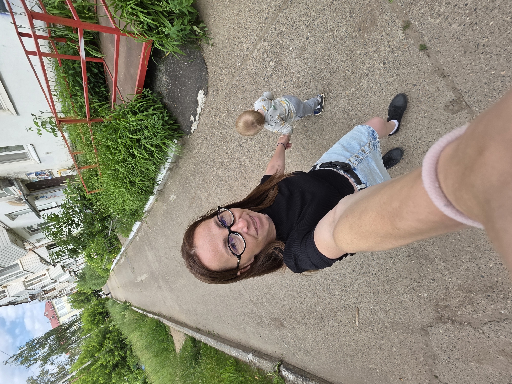 This is a first-person perspective selfie taken outdoors in Chaikovsky, Russia. The subject, a woman with long brown hair and glasses, is holding the hand of a small child who is walking on a concrete path. The child is wearing a grey long-sleeved shirt with a printed pattern and grey pants, along with dark shoes. The woman is wearing a black top, a denim skirt, and black sneakers. A pink band is visible on her upper arm.
The scene includes a sidewalk lined with green grass and some vegetation. To the left of the path, there is a building with white siding and windows, and a red railing leading up to what appears to be an entrance. The building suggests an urban or suburban residential setting. The sky is partly cloudy, indicating daytime with possibly mixed weather. The overall mood appears casual and perhaps joyful, as the woman is smiling at the camera while walking with the child. The concrete path shows some cracks and wear, typical of an outdoor public area. No visible text is present in the image.