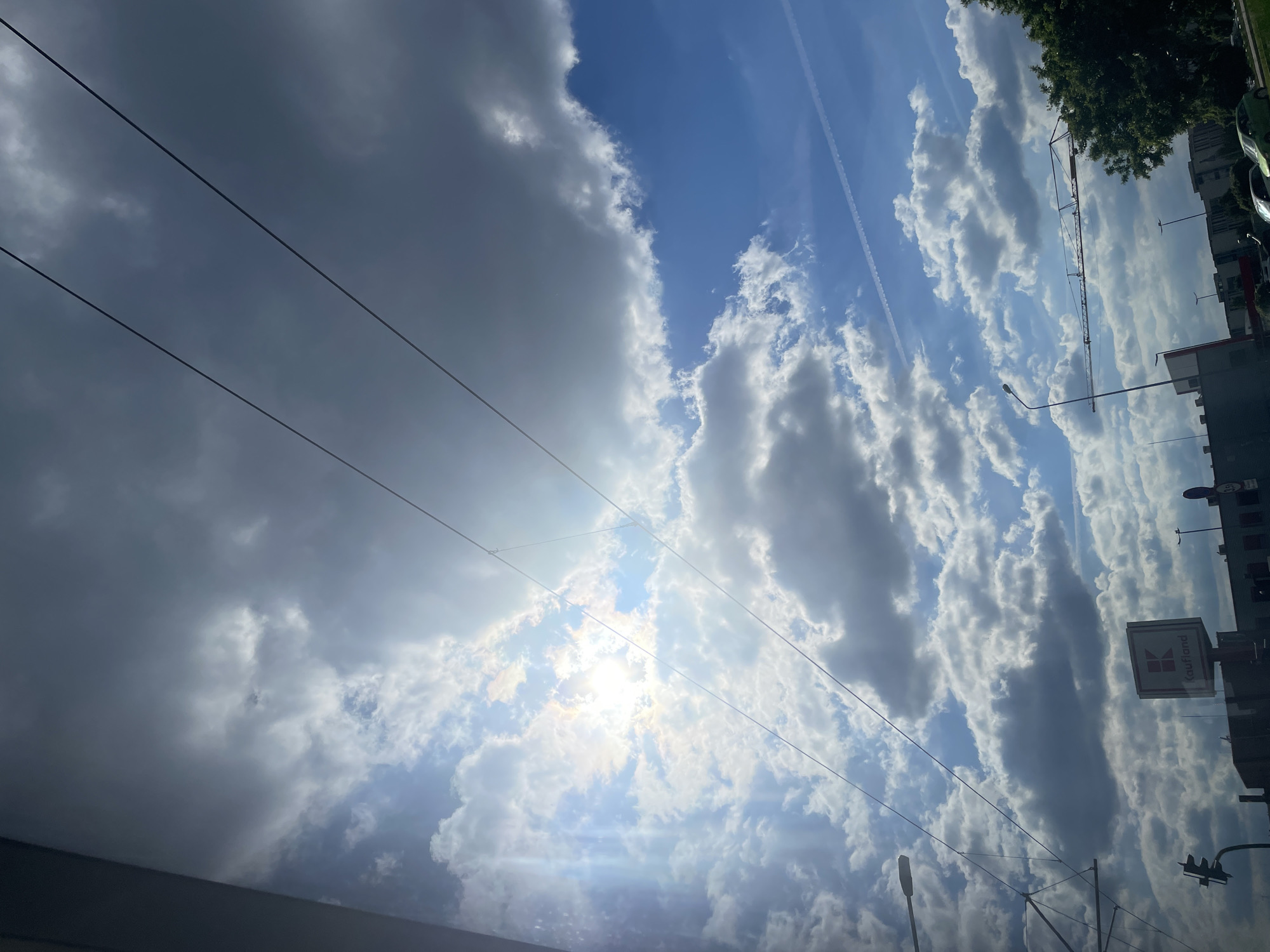 The image shows a bright sky with scattered clouds and a contrail, taken from a low angle in an urban environment. Overhead power lines are visible, extending across the sky. On the right side of the frame, blurred buildings, a traffic light, and a sign with "Kaufland" written on it are visible. Vegetation appears at the very top right corner. The exact location within the city of Unknown, Unknown cannot be determined.