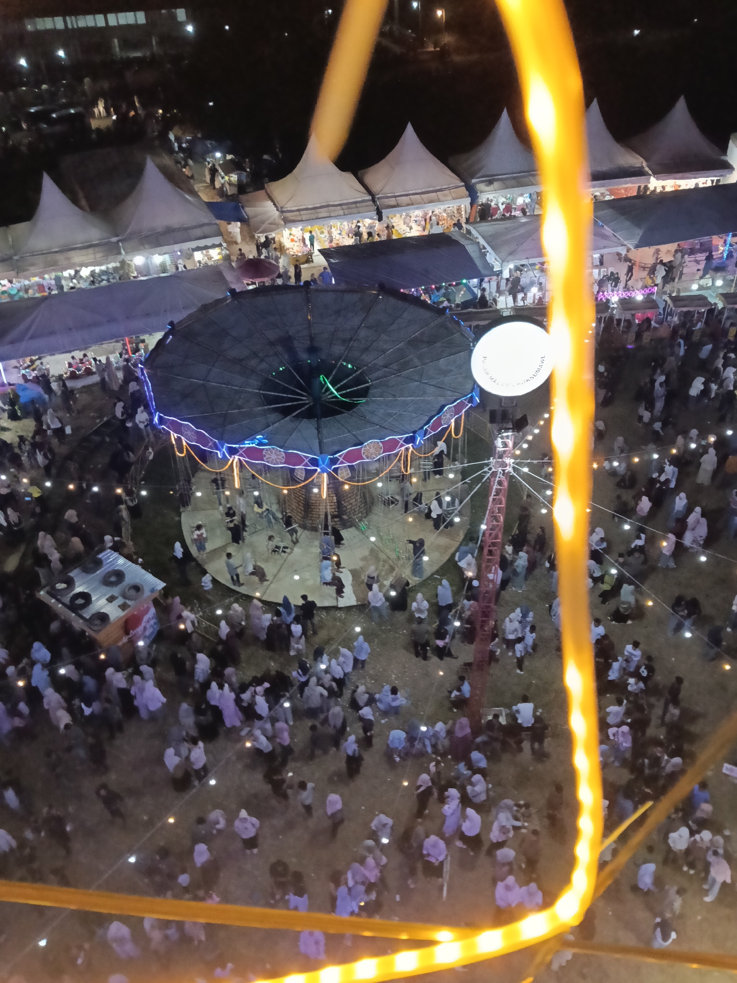 An aerial, night-time view captures a bustling outdoor fair in Keudepanteubreuh, Indonesia. A large, circular ride, adorned with colorful lights and a decorative border, is the central attraction. Numerous people are gathered around the ride and dispersed throughout the fairgrounds, which are lined with tents and stalls illuminated by strings of lights. The ground is a mix of dirt and grass. The image is partially obscured by a thick, glowing yellow element in the foreground, likely part of the viewpoint's structure, such as a Ferris wheel or similar amusement ride.