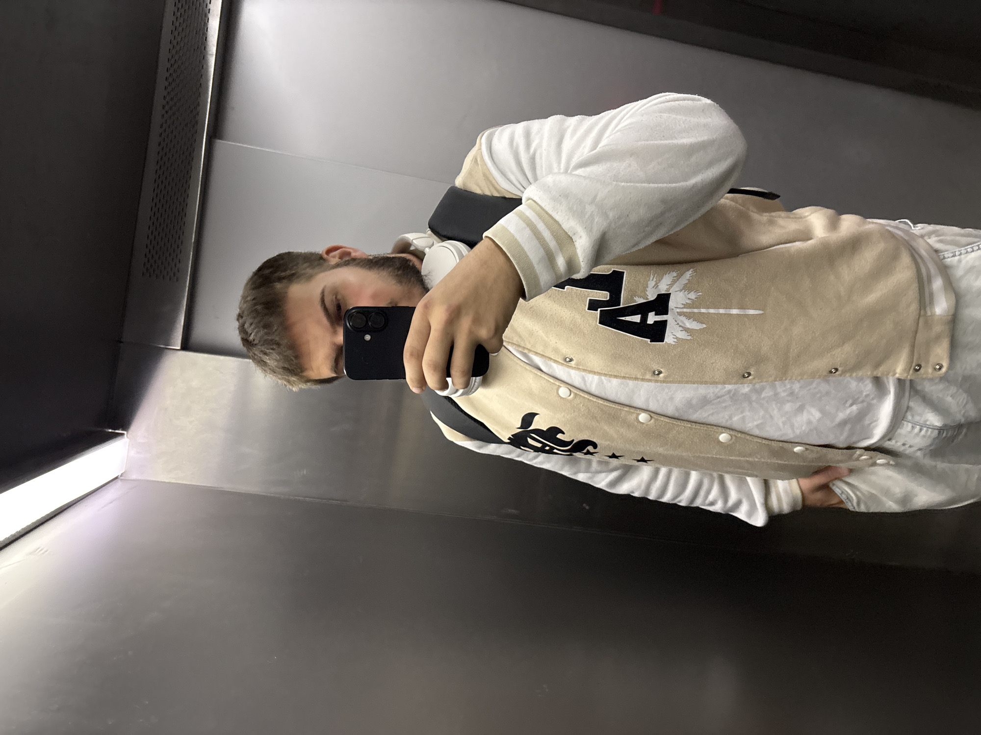 A person is taking a selfie in an elevator. They are wearing a white sweatshirt and a beige and white letterman jacket with black designs. They have headphones around their neck and are holding a black smartphone. The elevator has gray walls, an illuminated corner, and a dark ceiling. The location cannot be determined from the image.