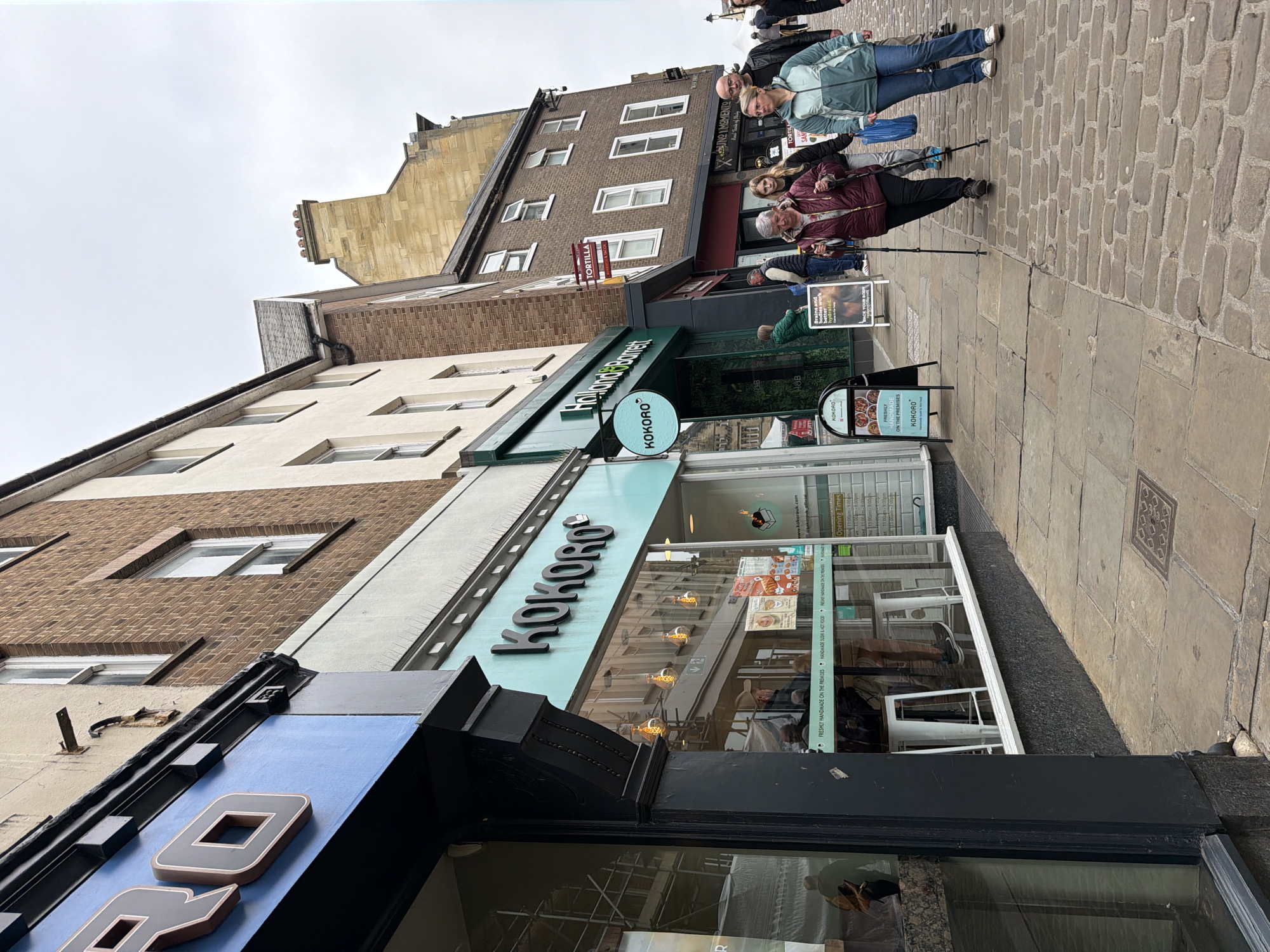 The image shows the exterior of a Kokoro restaurant located on a cobblestone street in the city of Durham, United Kingdom. Several people are walking past the restaurant on the sidewalk. The buildings surrounding the Kokoro have brick and stone facades. Signage for other businesses, including a Holland & Barrett and Tortilla, is visible. A sandwich board displays the message "FRESHLY HANDMADE ON THE PREMISES". Opening times for Kokoro are displayed on another A-board. The sky is overcast.